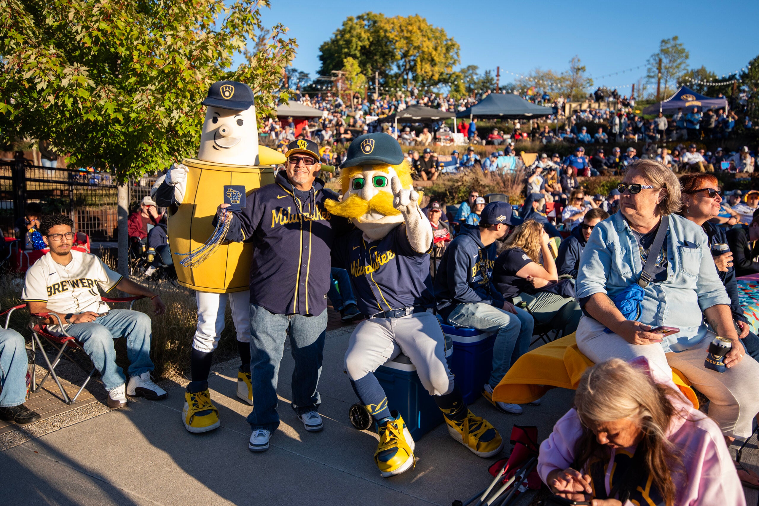 A crowd watches an outdoor event featuring two mascots, one dressed as a barrel and the other as a bird, with people sitting and standing around them in casual attire.