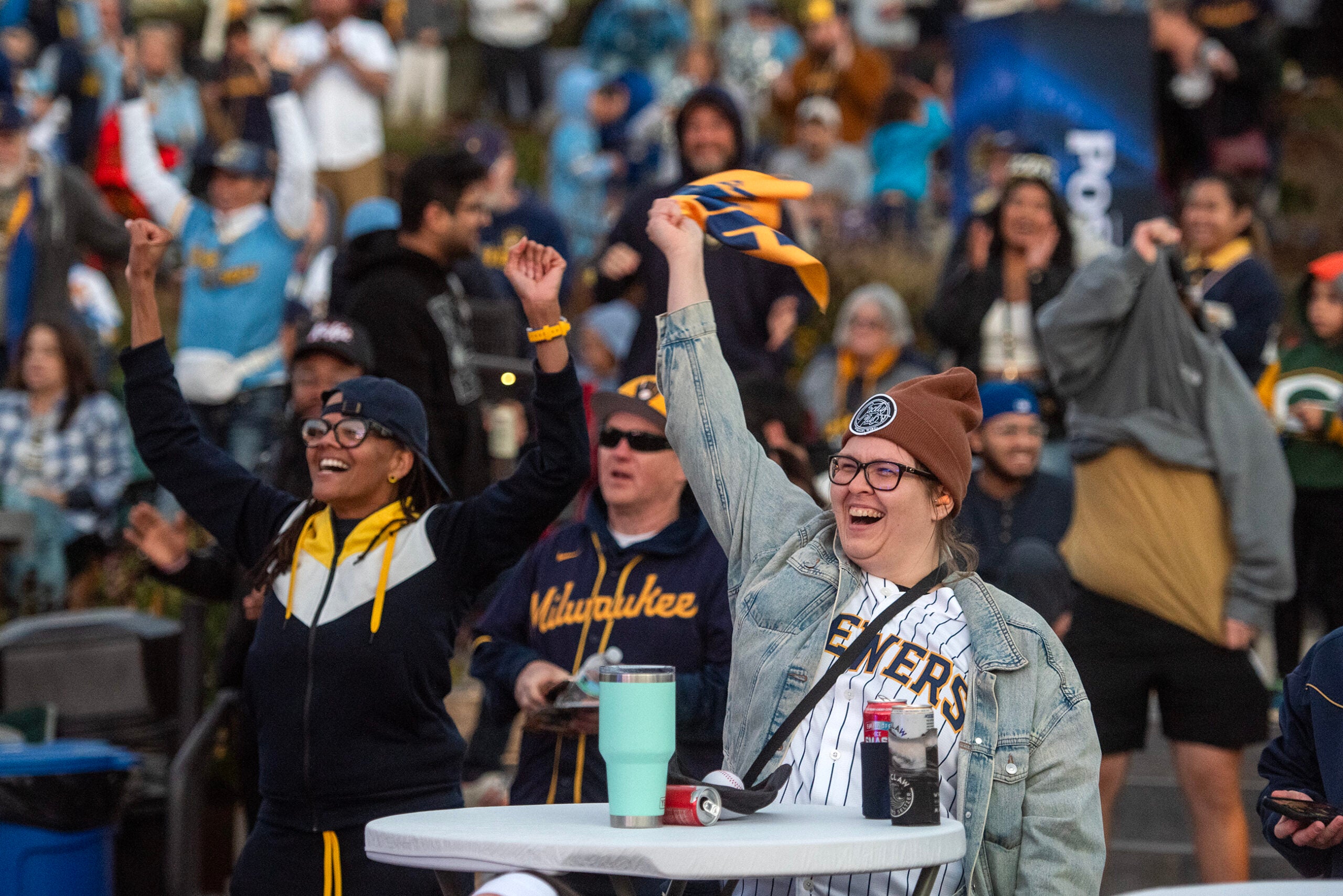 Two women cheer with raised arms at an outdoor sporting event, surrounded by a crowd of enthusiastic fans in team apparel.