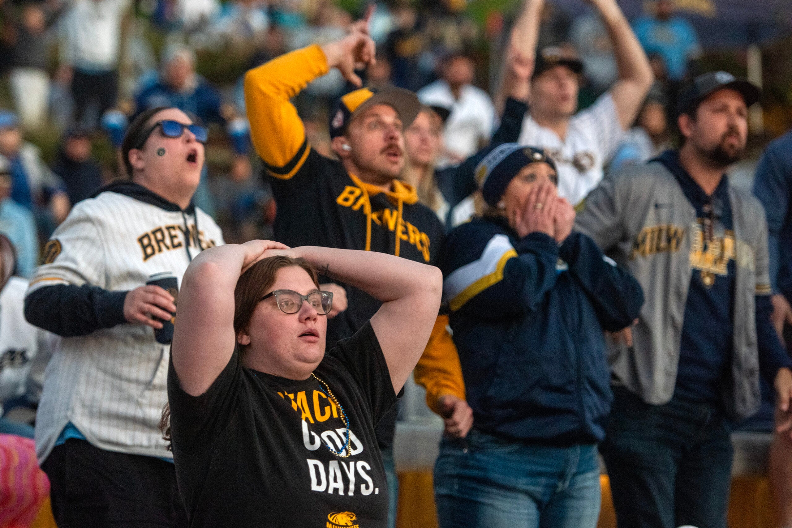 A group of Milwaukee Brewers fans in team apparel react emotionally while watching a game, with some holding their heads in disbelief.