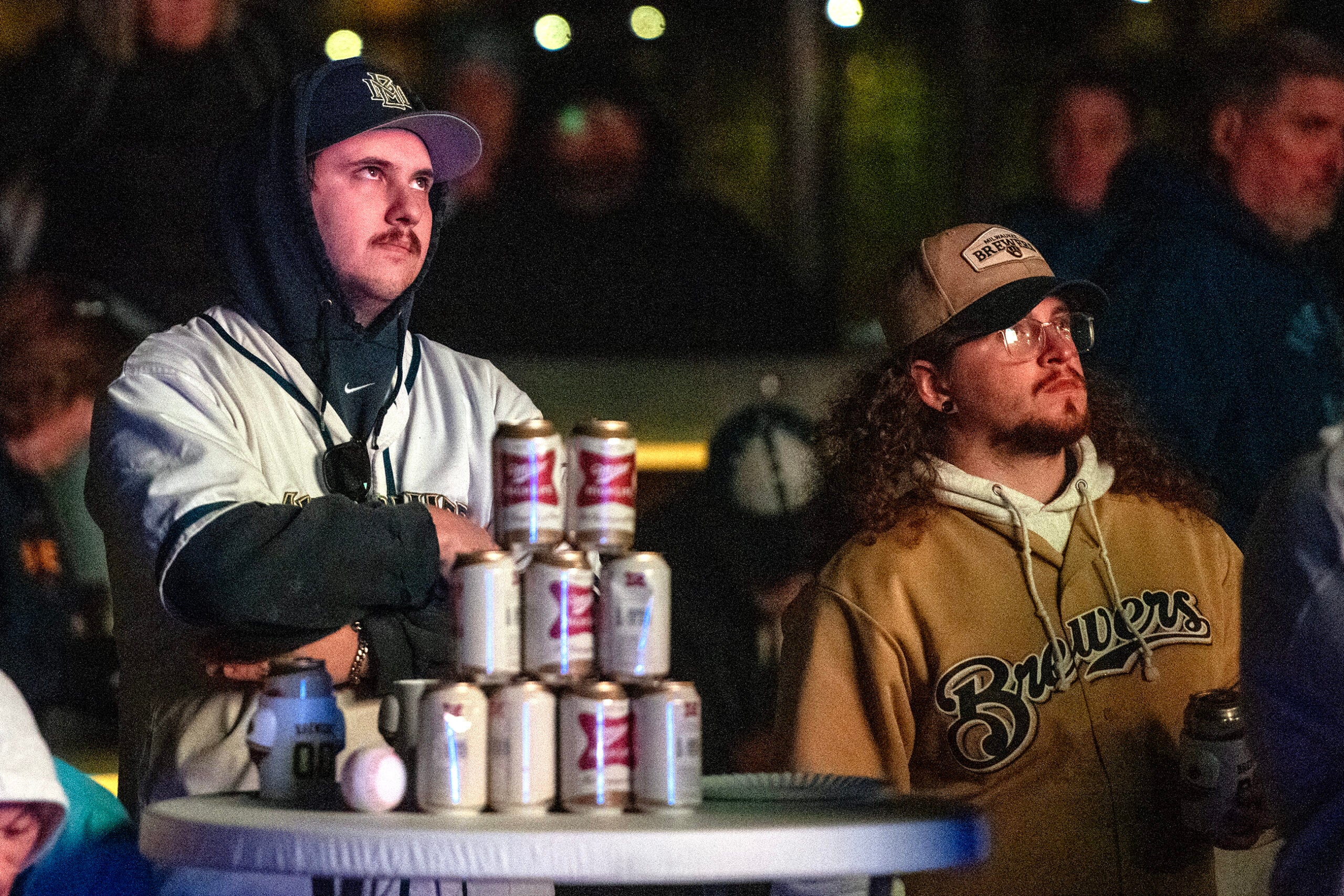 Two men in baseball attire stand behind a table stacked with cans of beer and a baseball, watching something intently with a crowd in the background.