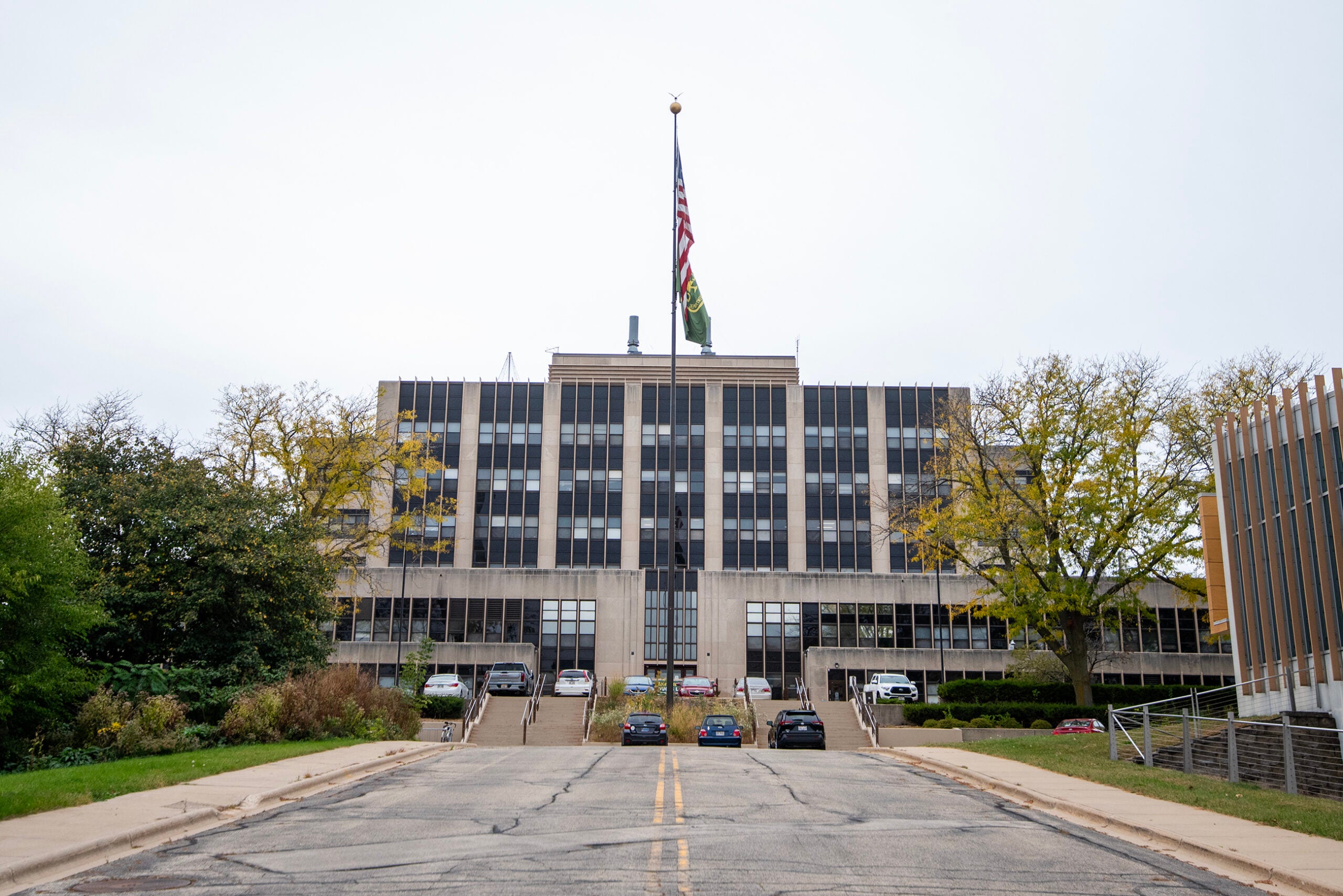 A government building with multiple floors, an American flag on the roof, several cars parked in front, and trees on either side of the entrance.