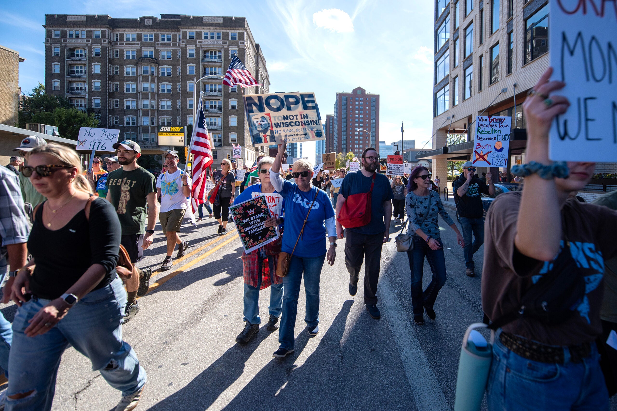 A group of people march down a city street holding protest signs, including one reading NOPE NOT IN WISCONSIN, with buildings visible in the background.