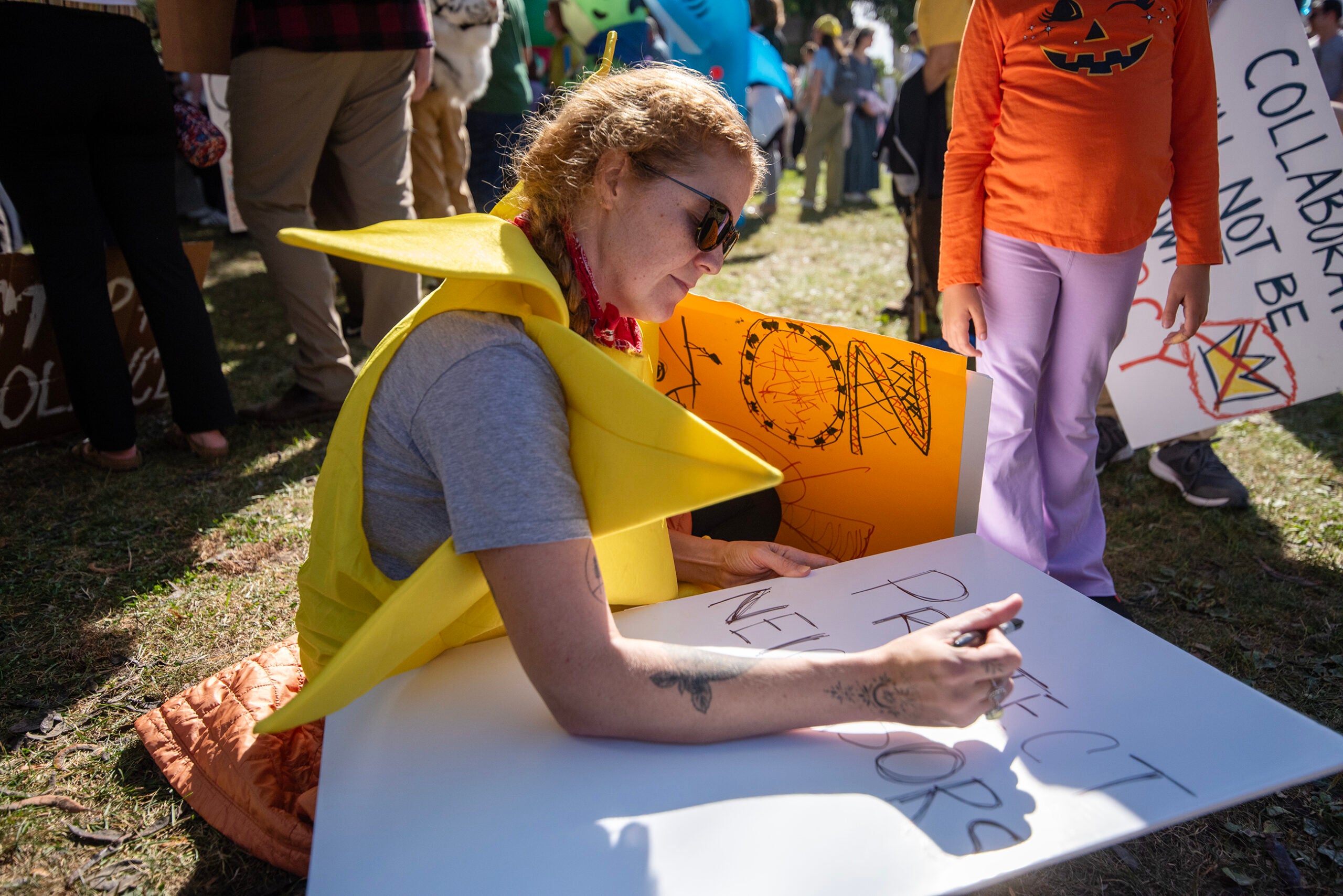 A person wearing a yellow costume sits on the grass and writes “PROTECT NEIGHBORS” on a sign at an outdoor gathering. Other people holding signs are visible in the background.