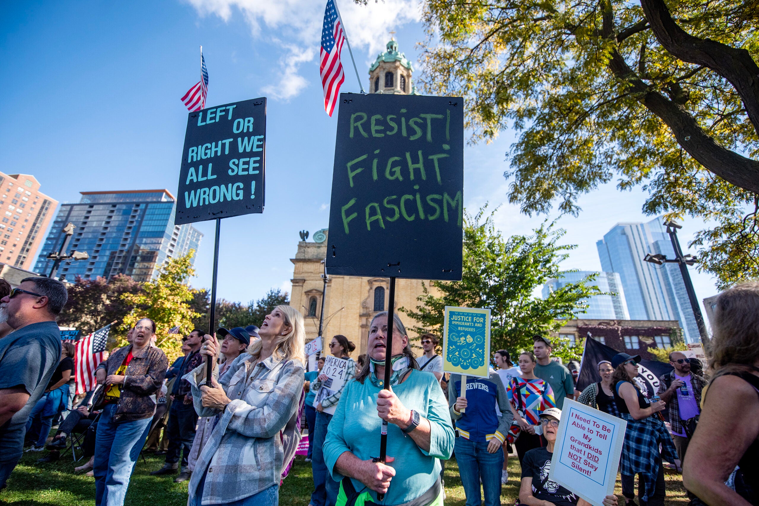 A group of protesters gather outdoors, some holding signs that read LEFT OR RIGHT WE ALL SEE WRONG! and RESIST! FIGHT FASCISM. Buildings and trees are visible in the background.