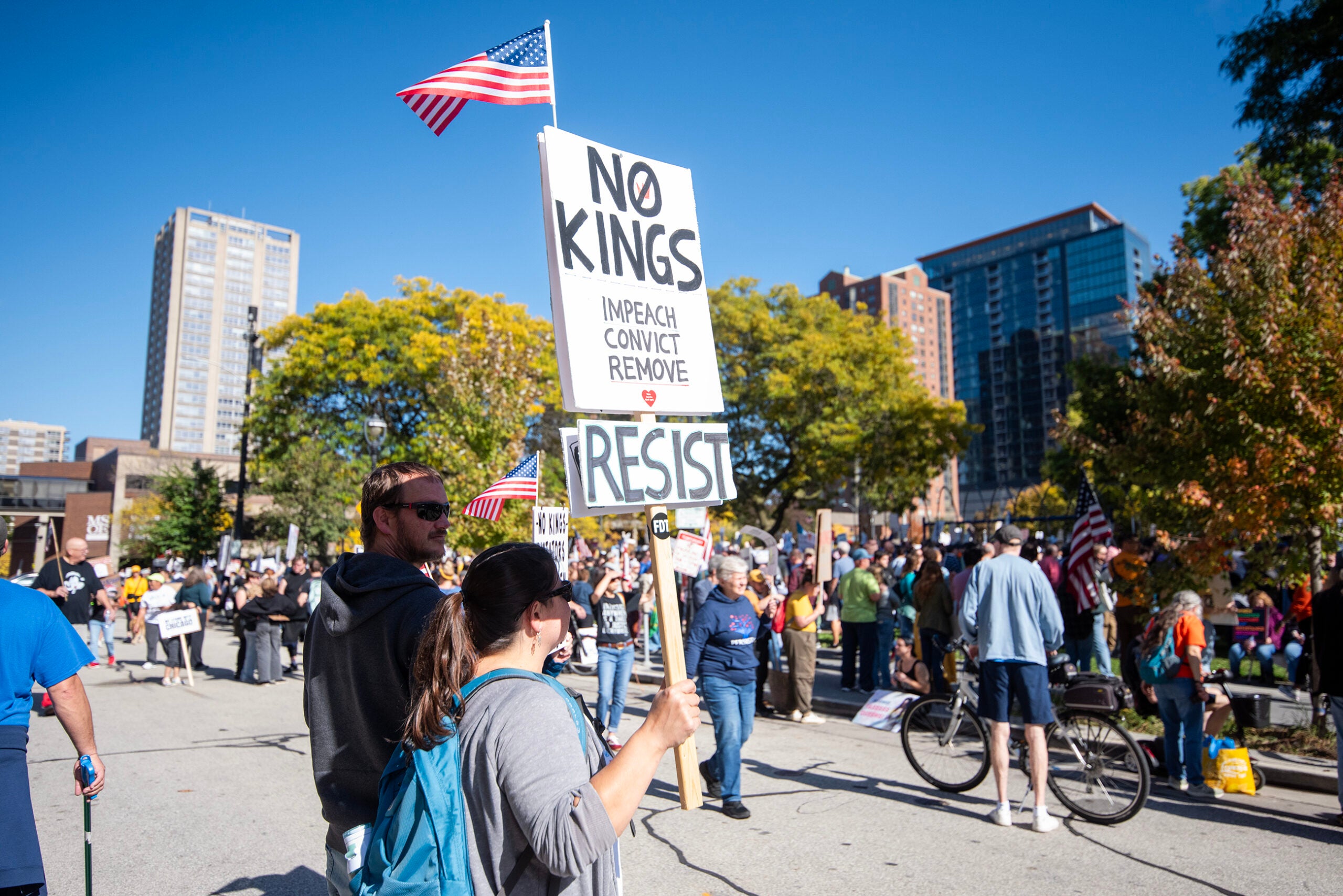 Protesters hold signs reading NO KINGS, IMPEACH CONVICT REMOVE, and RESIST at a street demonstration with American flags and city buildings in the background.