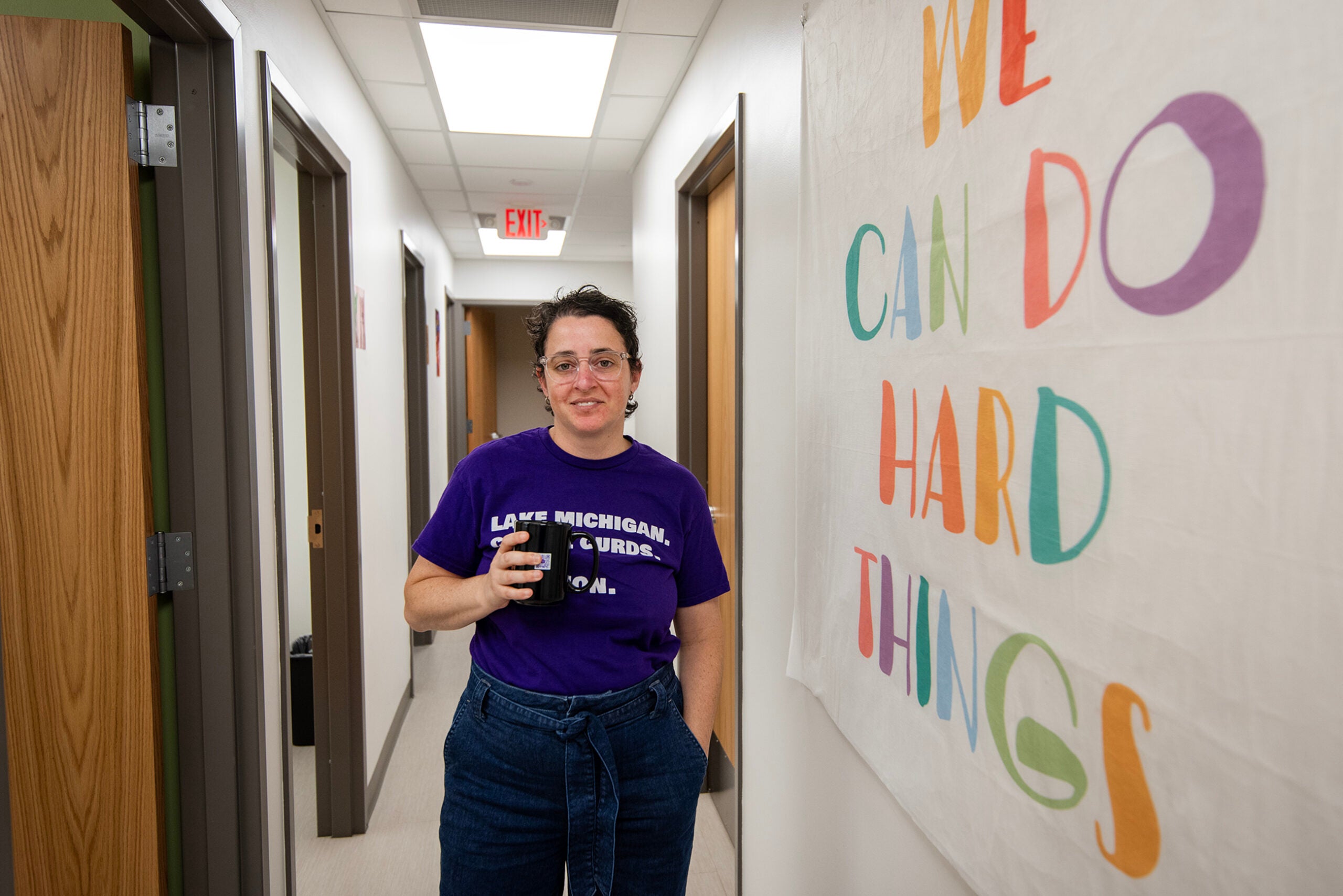 A person in glasses stands in a hallway holding a mug, next to a wall sign that reads “We can do hard things.”.