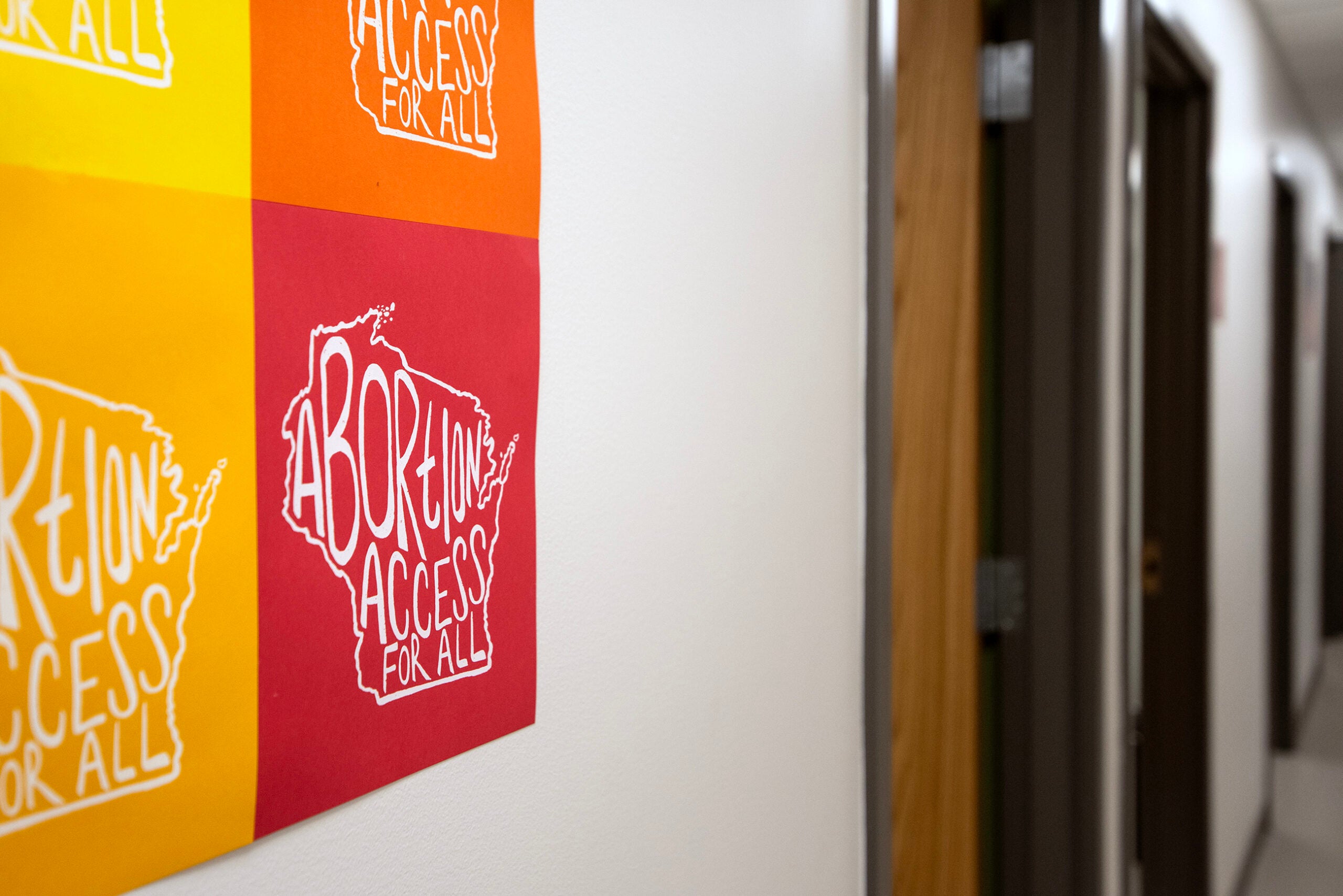 A poster on a hallway wall features the phrase Abortion Access For All in bold text on red, orange, and yellow backgrounds.