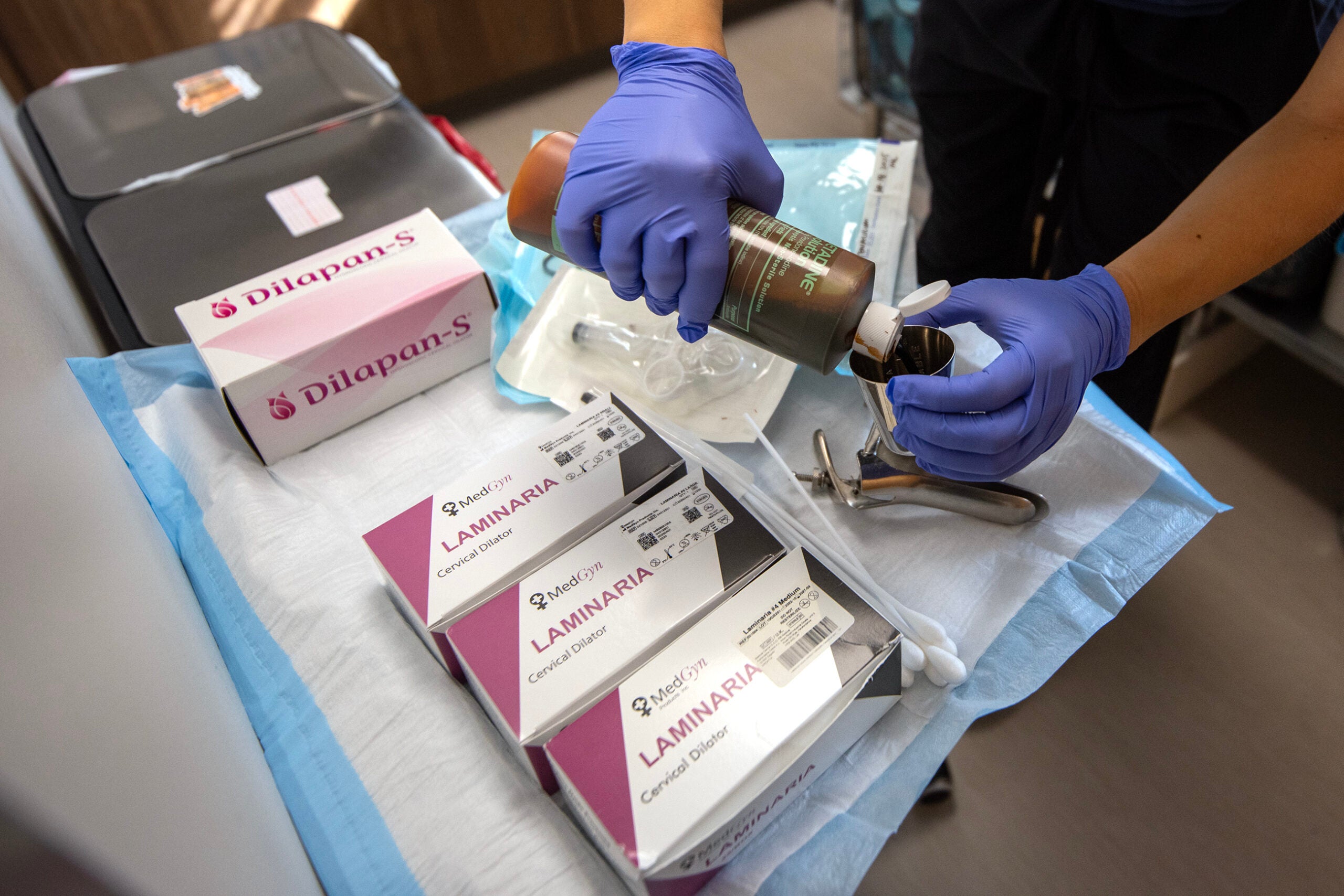 A person wearing blue gloves prepares medical instruments and supplies, including Dilapan-S and Laminaria cervical dilators, on a covered table.