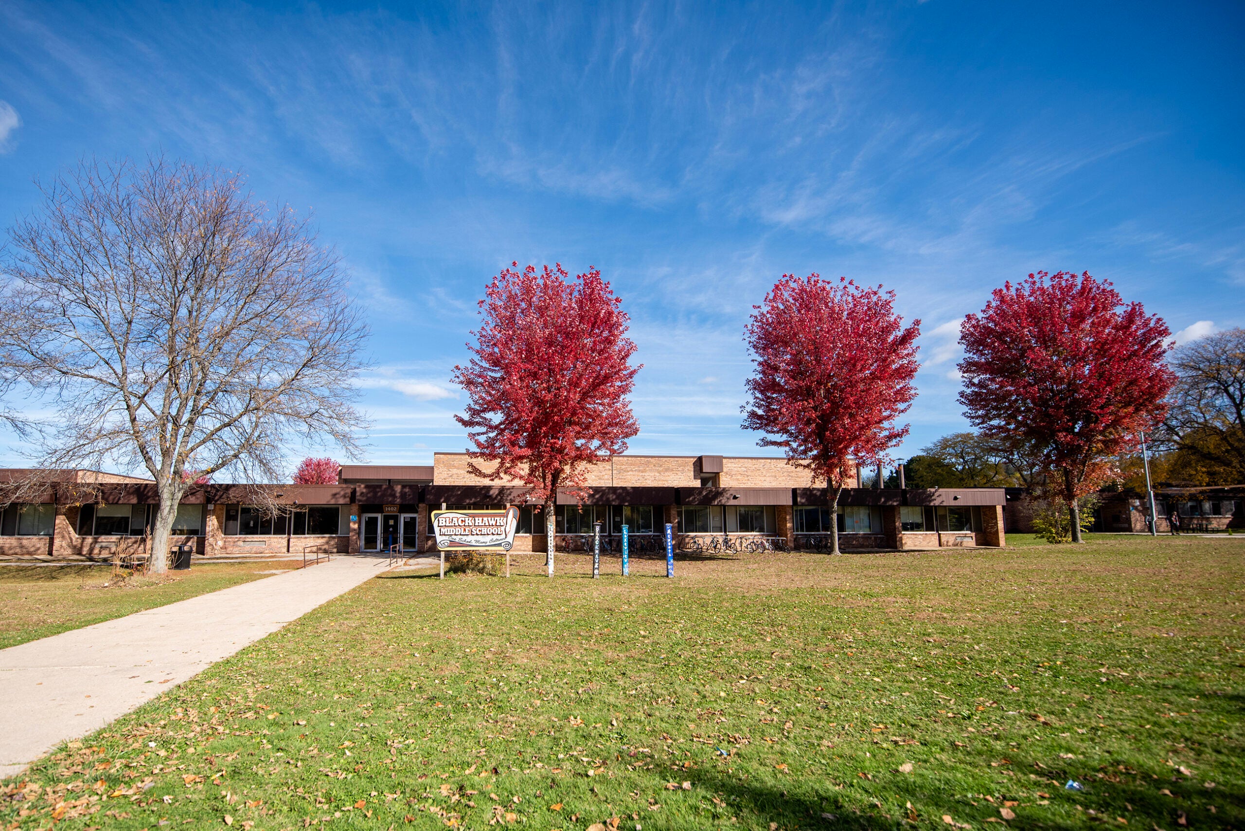A single-story school building with a brown exterior, a front lawn, four red-leaved trees, a sidewalk, and a wooden sign under a blue sky.