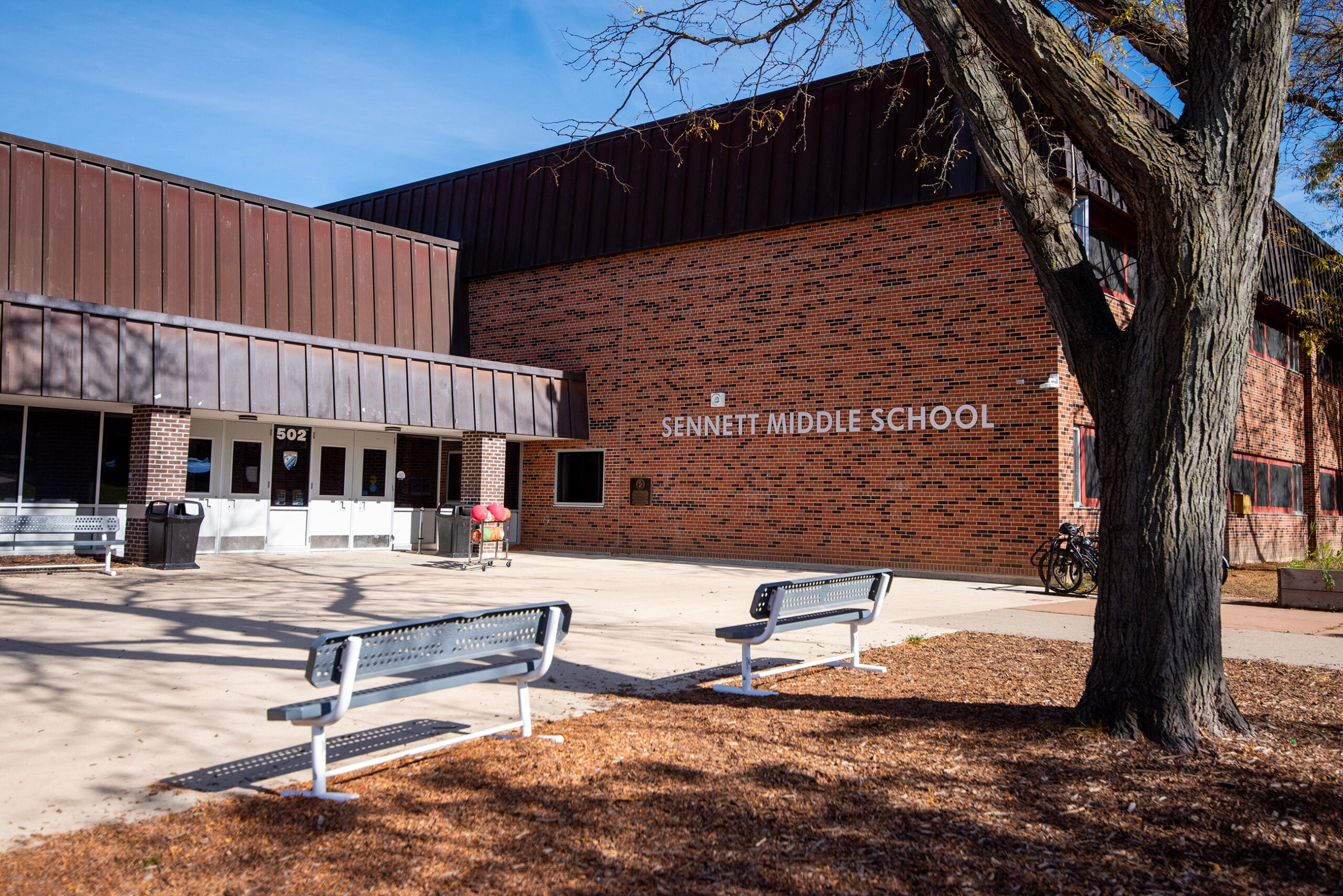 Exterior view of Sennett Middle School, showing brick building, entrance doors, benches, tree, and two bicycles near the wall.