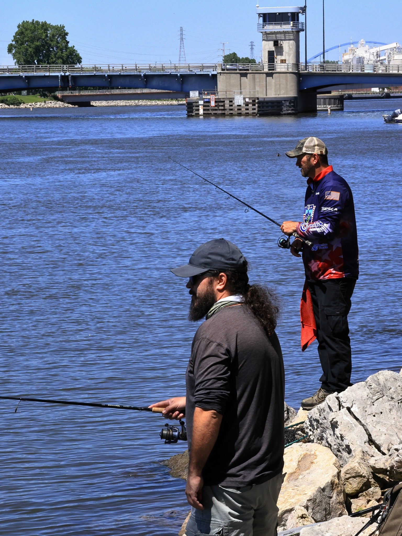 Two men stand on rocky shore fishing in a river, with a bridge and control tower visible in the background on a sunny day.