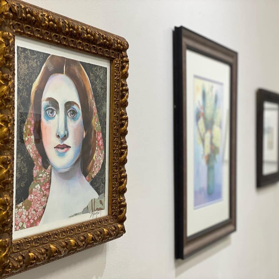 Framed paintings hang on a white wall; the closest features a stylized portrait of a woman, while the others display floral artwork.
