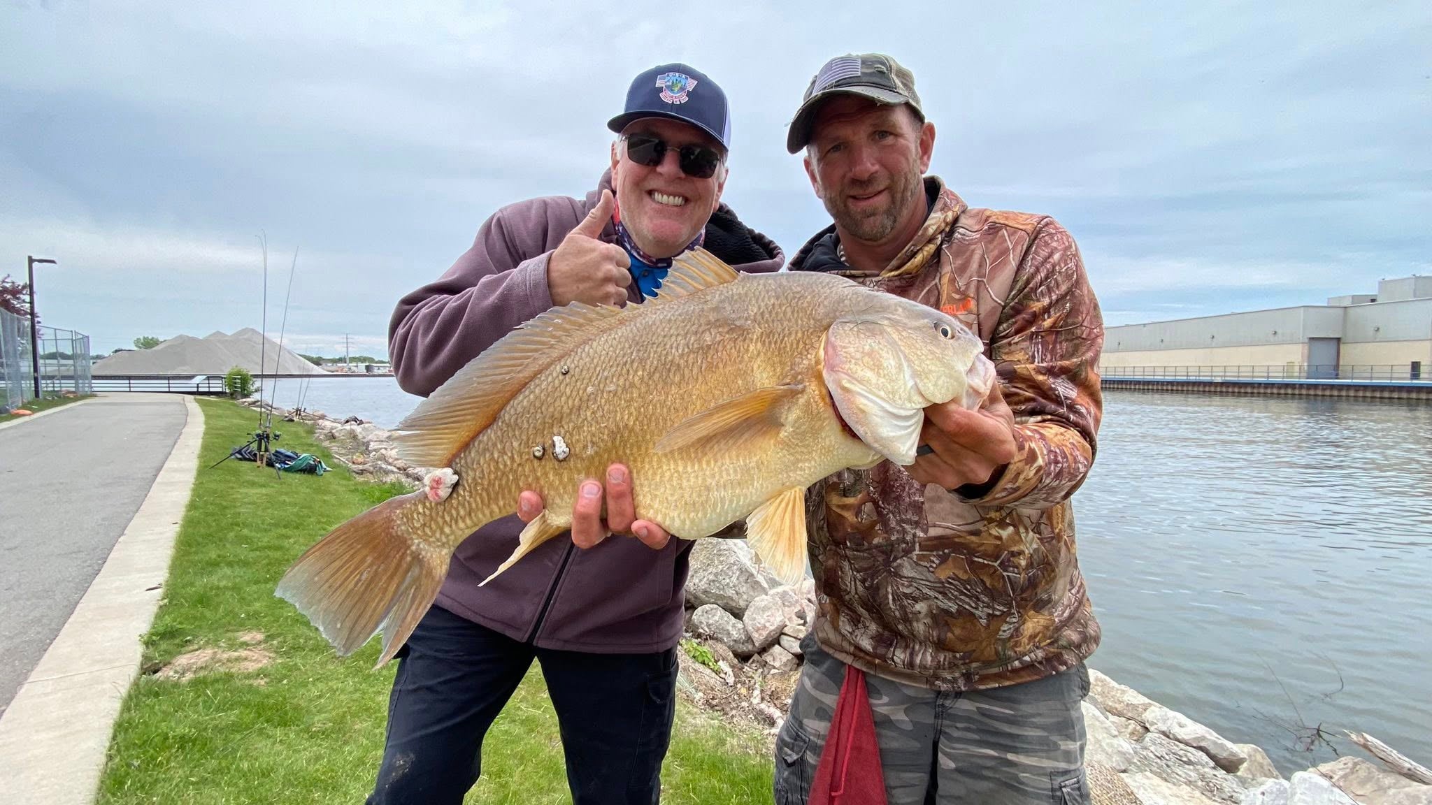 Two men standing by the water, smiling and holding a large fish. One gives a thumbs up. Industrial buildings and a grassy area are visible in the background.