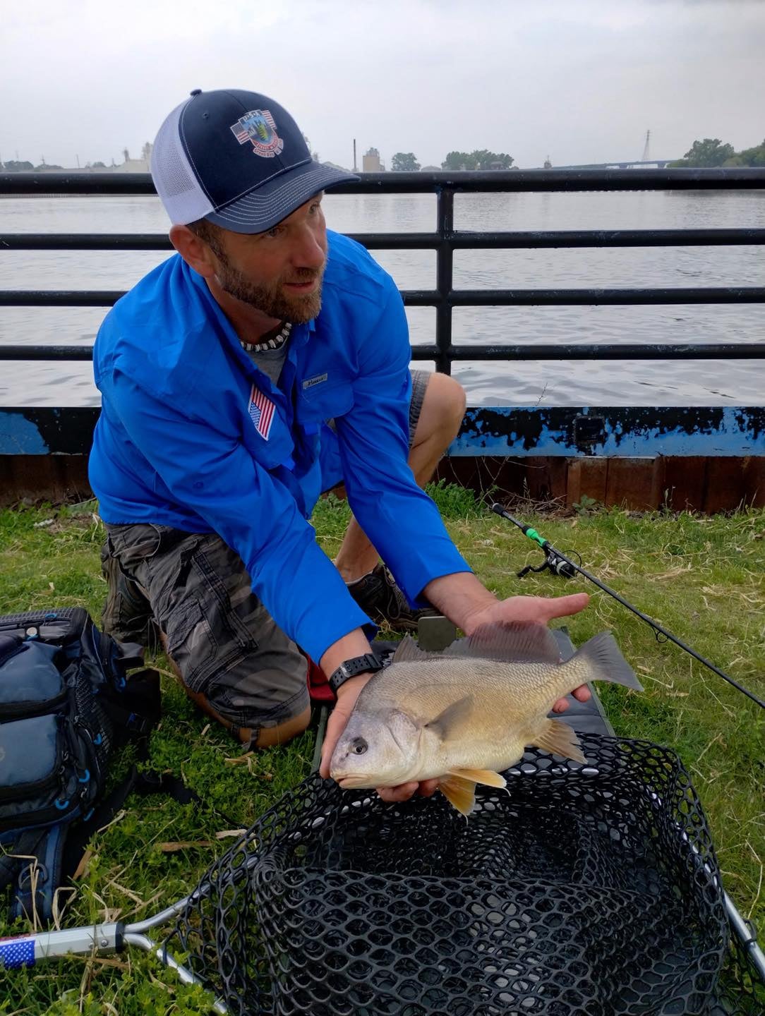 A man in a blue shirt and cap kneels by a river, holding a large fish over a net on the grass, with fishing gear nearby.
