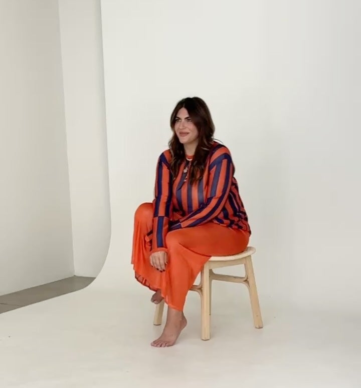 A woman with long brown hair sits barefoot on a wooden stool, wearing an orange and navy striped sweater and an orange skirt, in a minimalist studio setting with a light background.