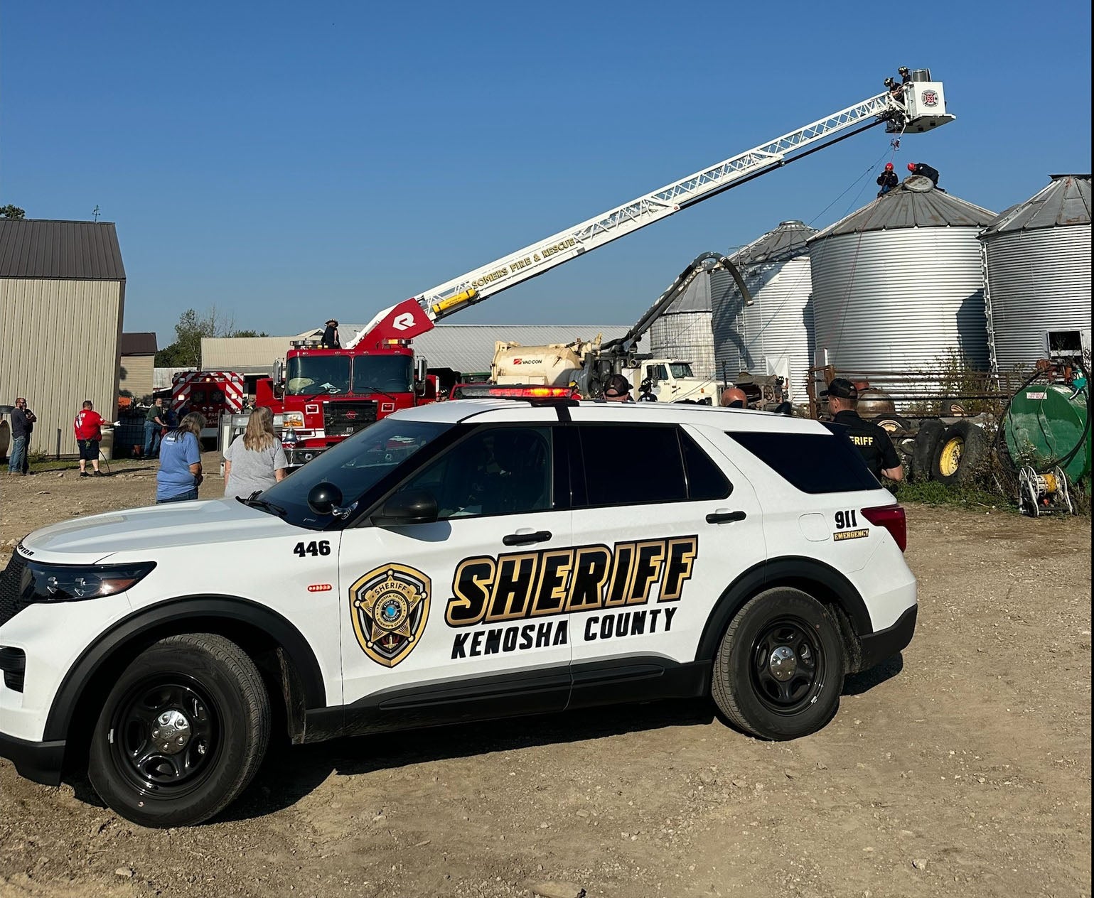 A Kenosha County Sheriff SUV is parked in front of fire trucks and silos as emergency responders work on top of a grain bin. Several people are observing the scene.