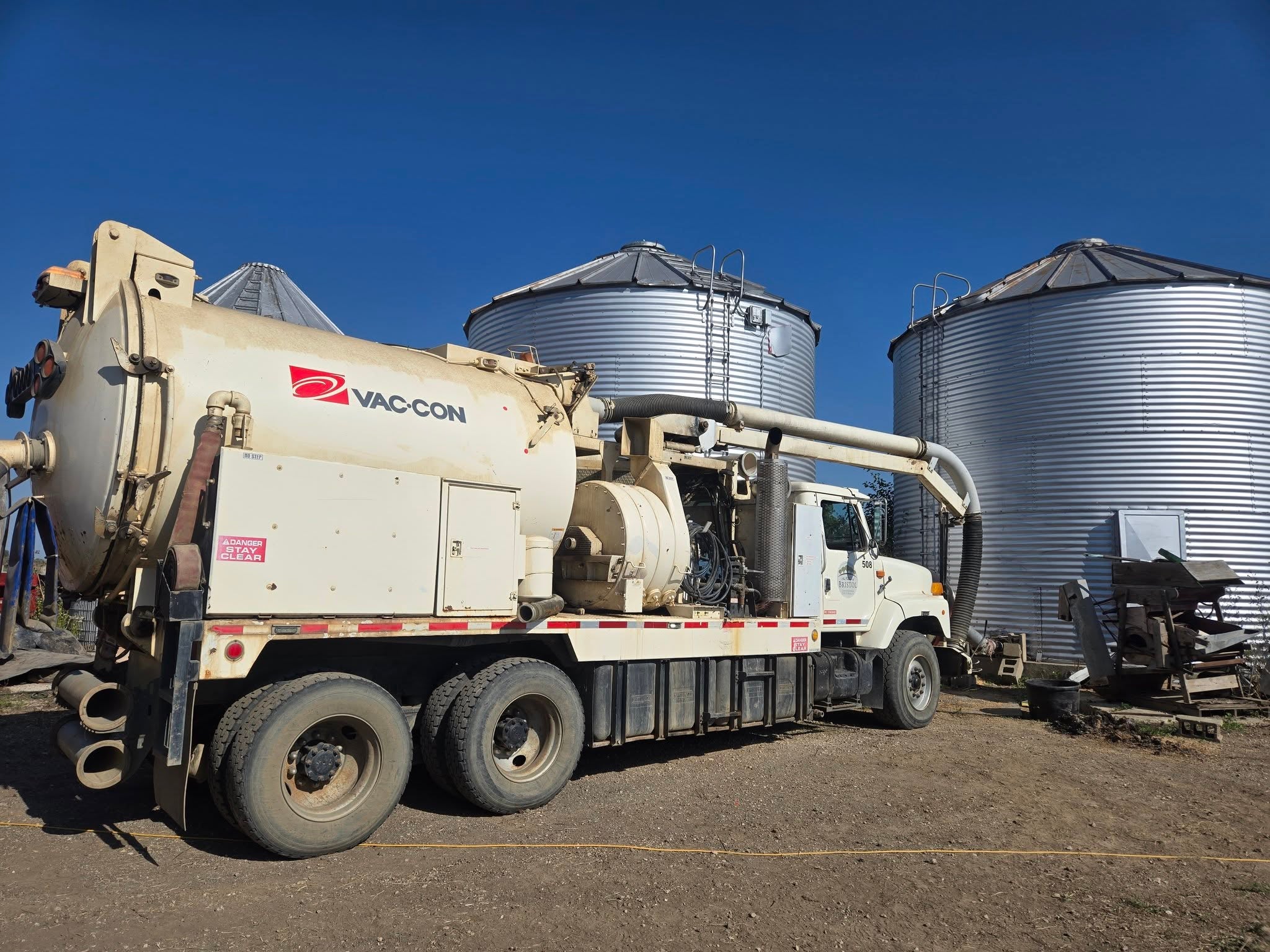 A Vac-Con vacuum truck is parked on dirt near two large metal grain silos under a clear blue sky.