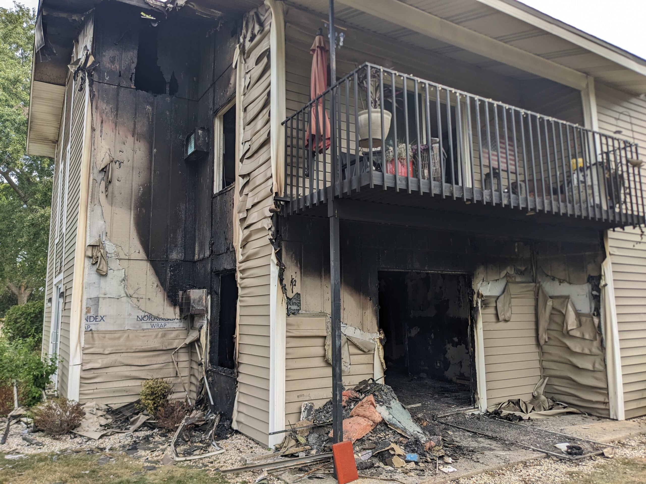 Two-story building with extensive fire damage; siding is melted and blackened, windows broken, and debris scattered on the ground outside.