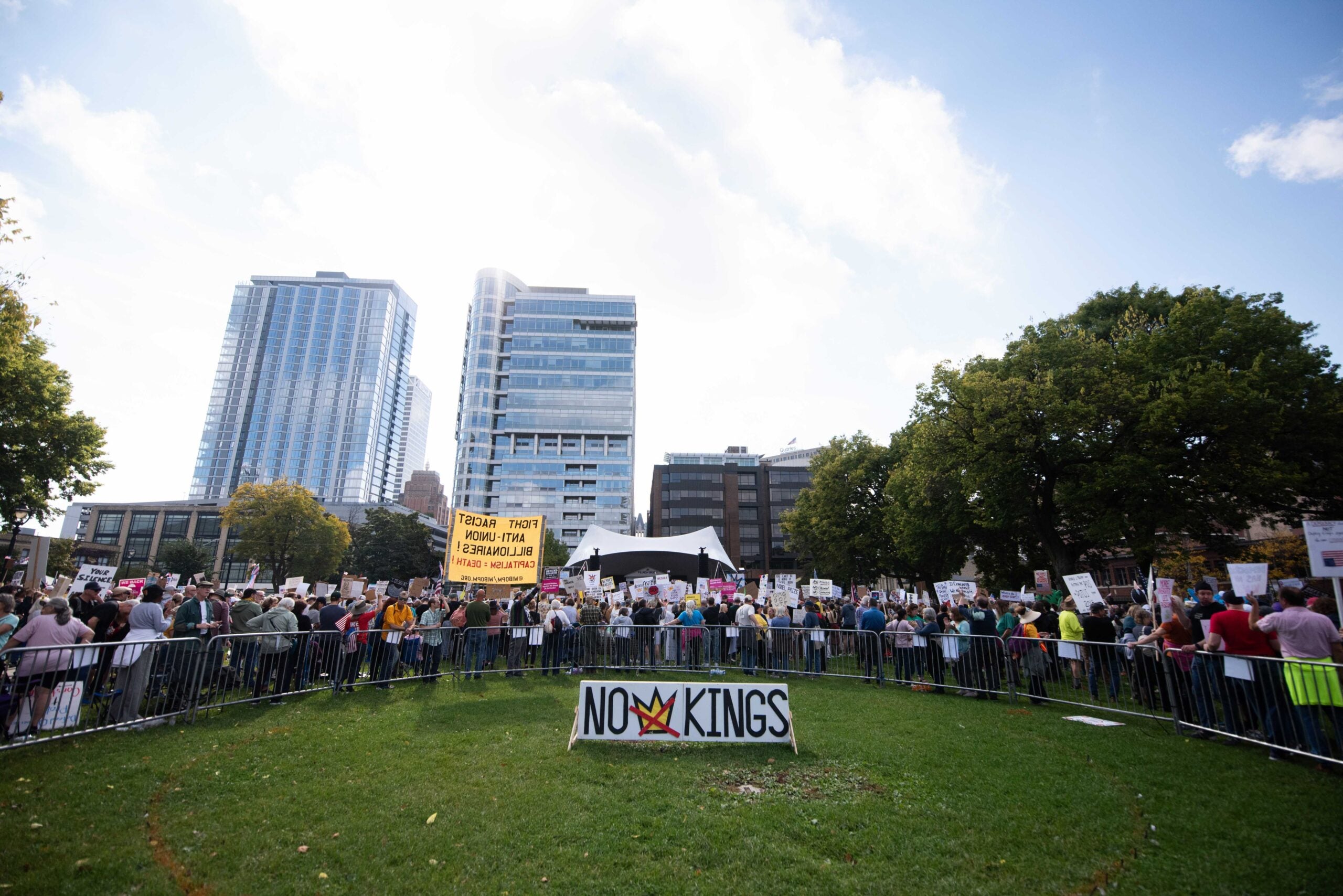 A crowd of people holding protest signs gathers in a park surrounded by tall buildings; a banner in front reads NO KINGS.