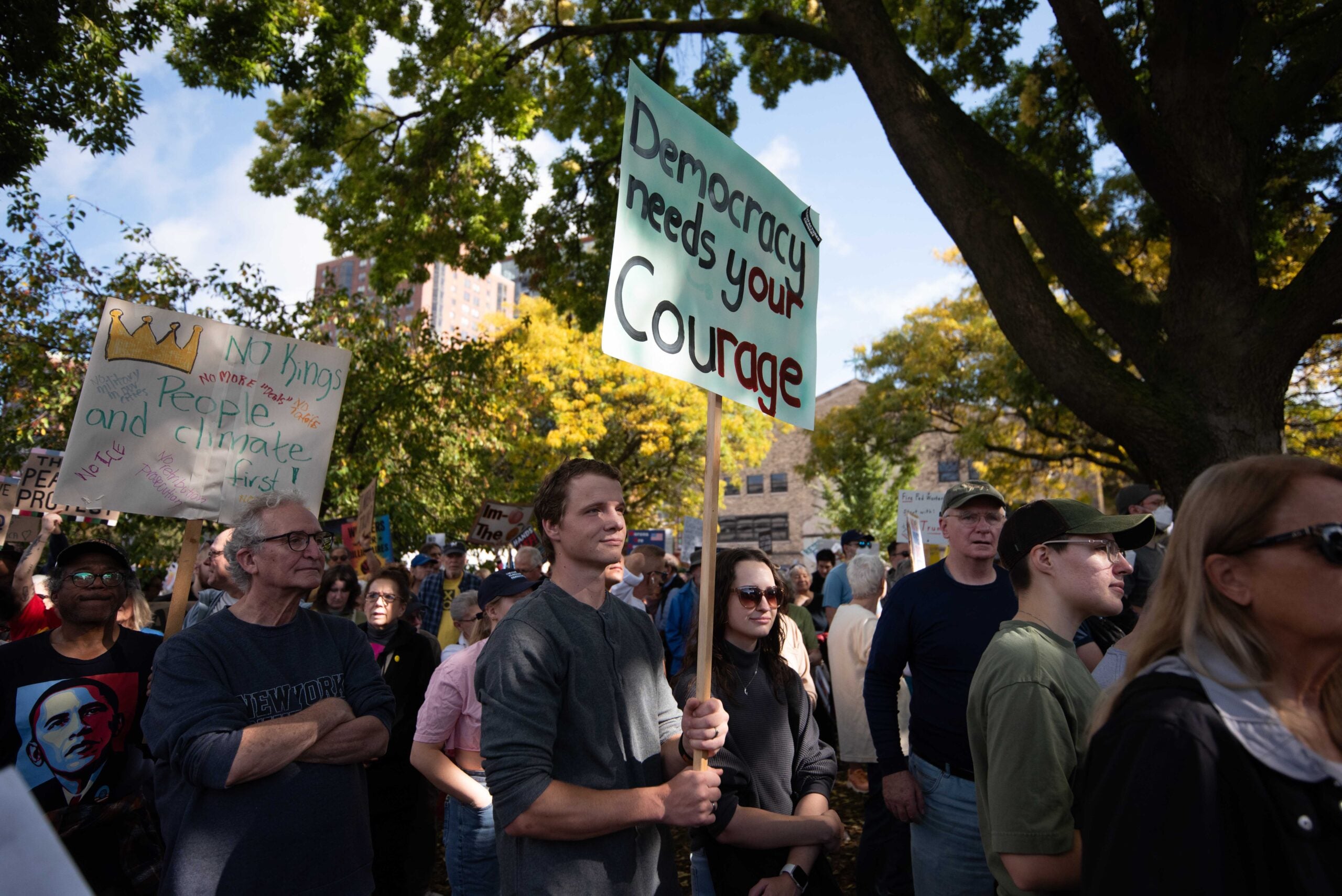 A group of people gathered outdoors at a protest; one person holds a sign reading, Democracy needs your Courage.