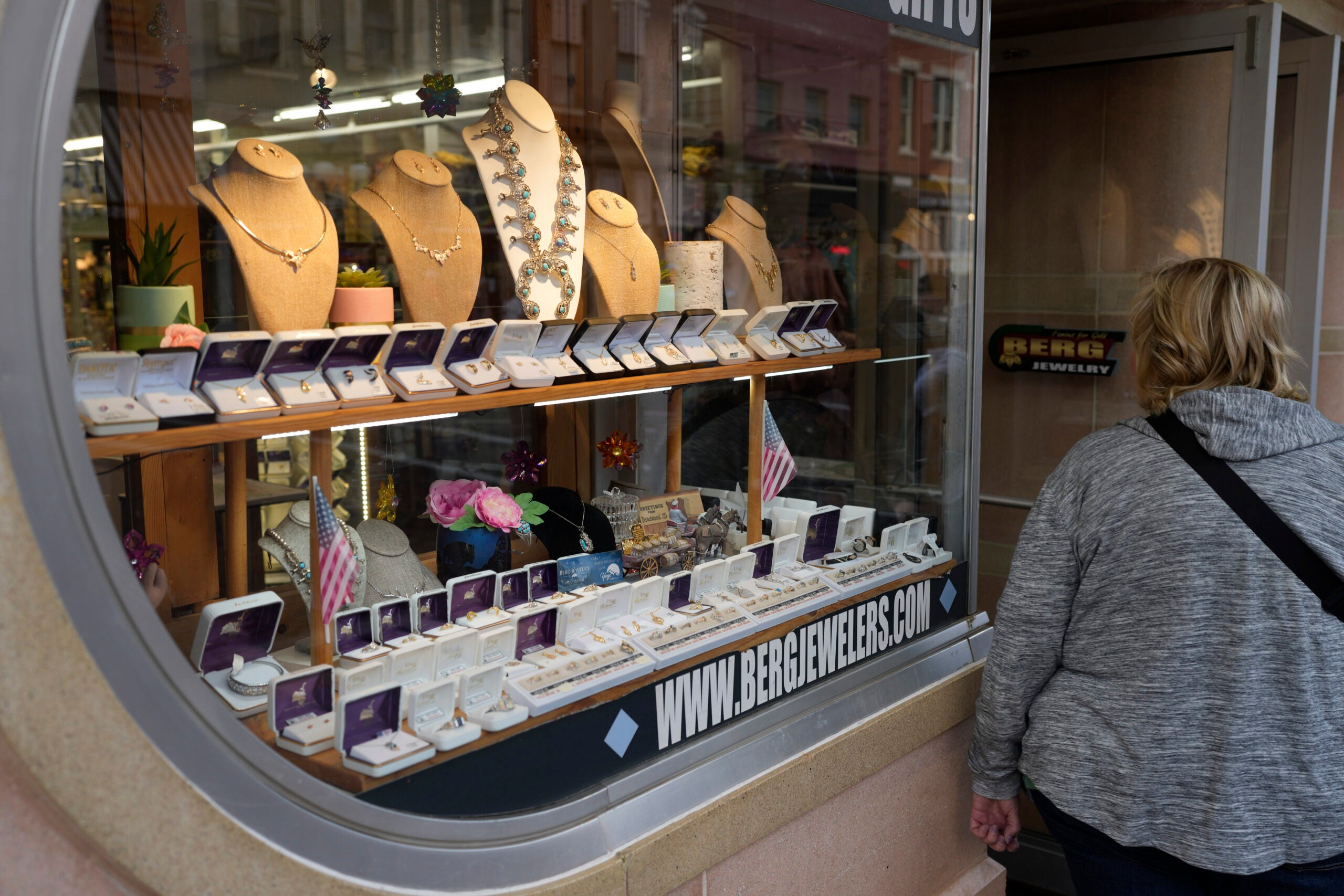 A person looks into a jewelry store window displaying necklaces, rings, and other jewelry on stands and in cases.