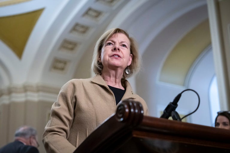 A woman in a tan coat stands at a podium with a microphone in a building with arched ceilings, addressing an audience.