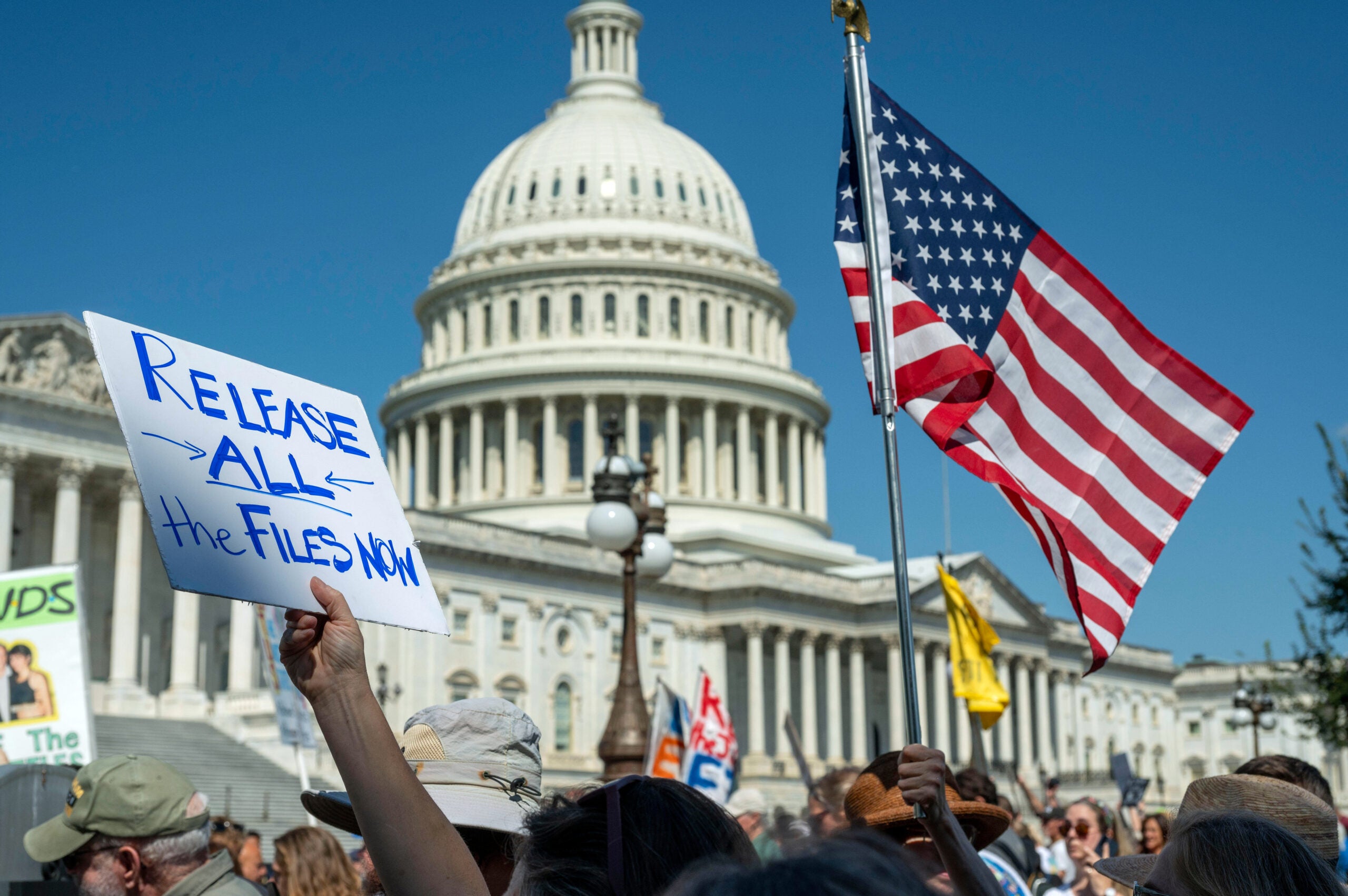 A crowd gathers in front of the U.S. Capitol, holding protest signs and an American flag; one sign reads, Release all the files now.