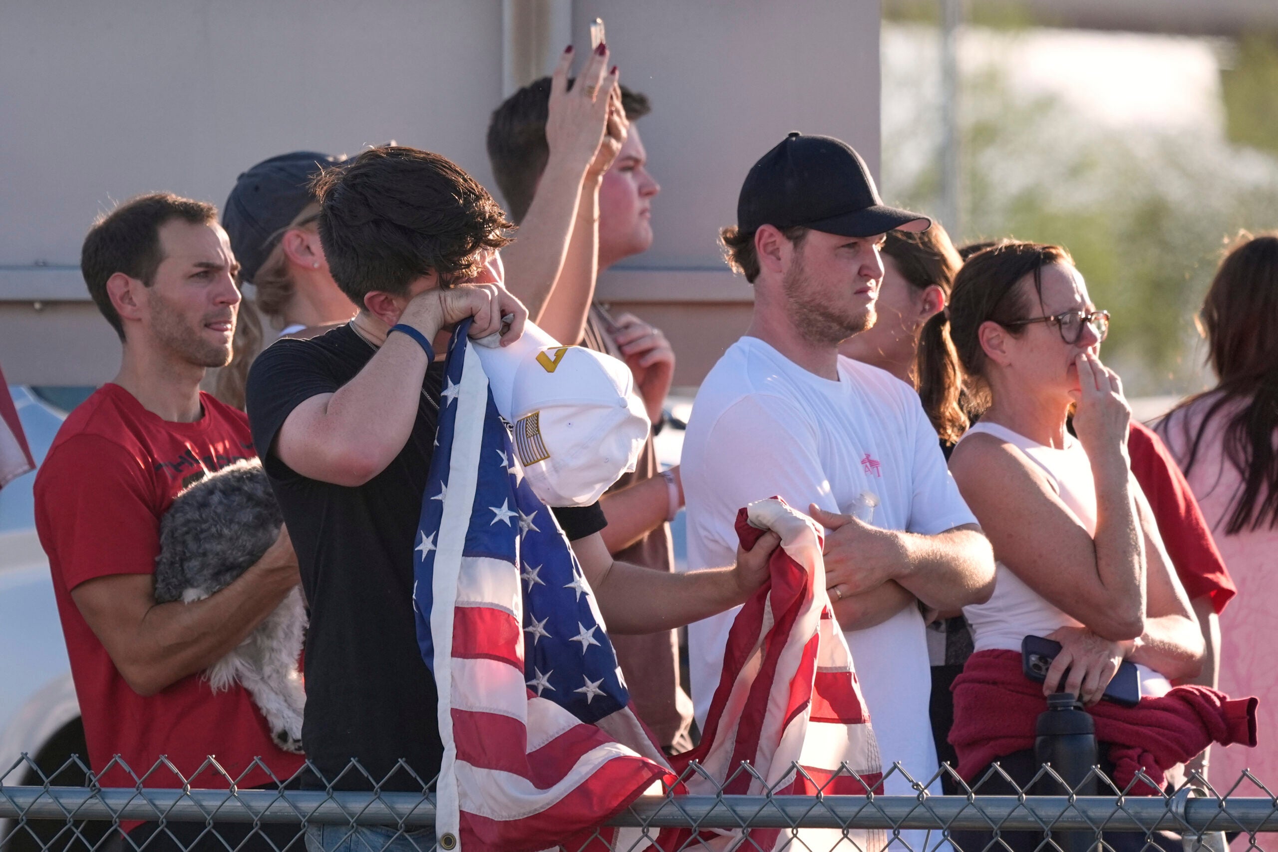 A group of people stand behind a fence, some appearing emotional, while one person wipes their face and holds a folded American flag.