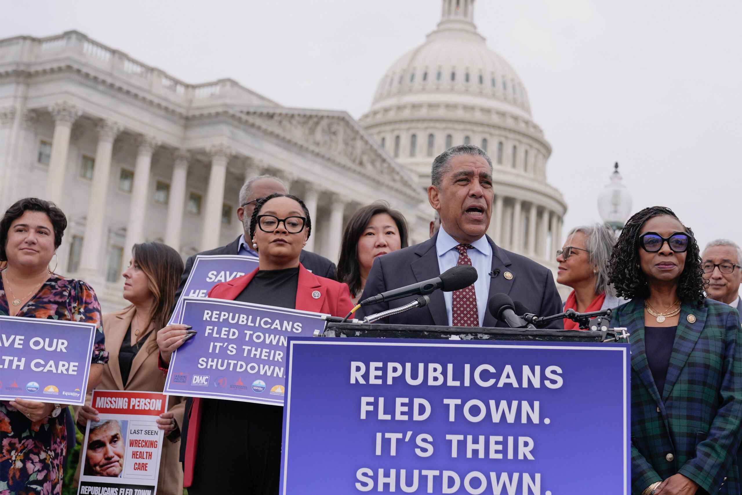 A group of people stand in front of the U.S. Capitol holding signs that read Republicans fled town. It’s their shutdown, as a man speaks at a podium with the same message.