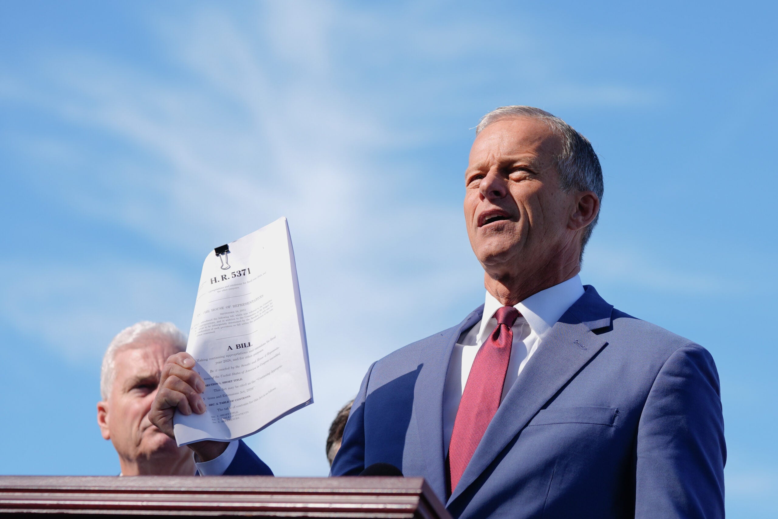 A man in a blue suit and red tie speaks at a podium outdoors, holding a printed document labeled H.R. 5741 with others standing behind him.