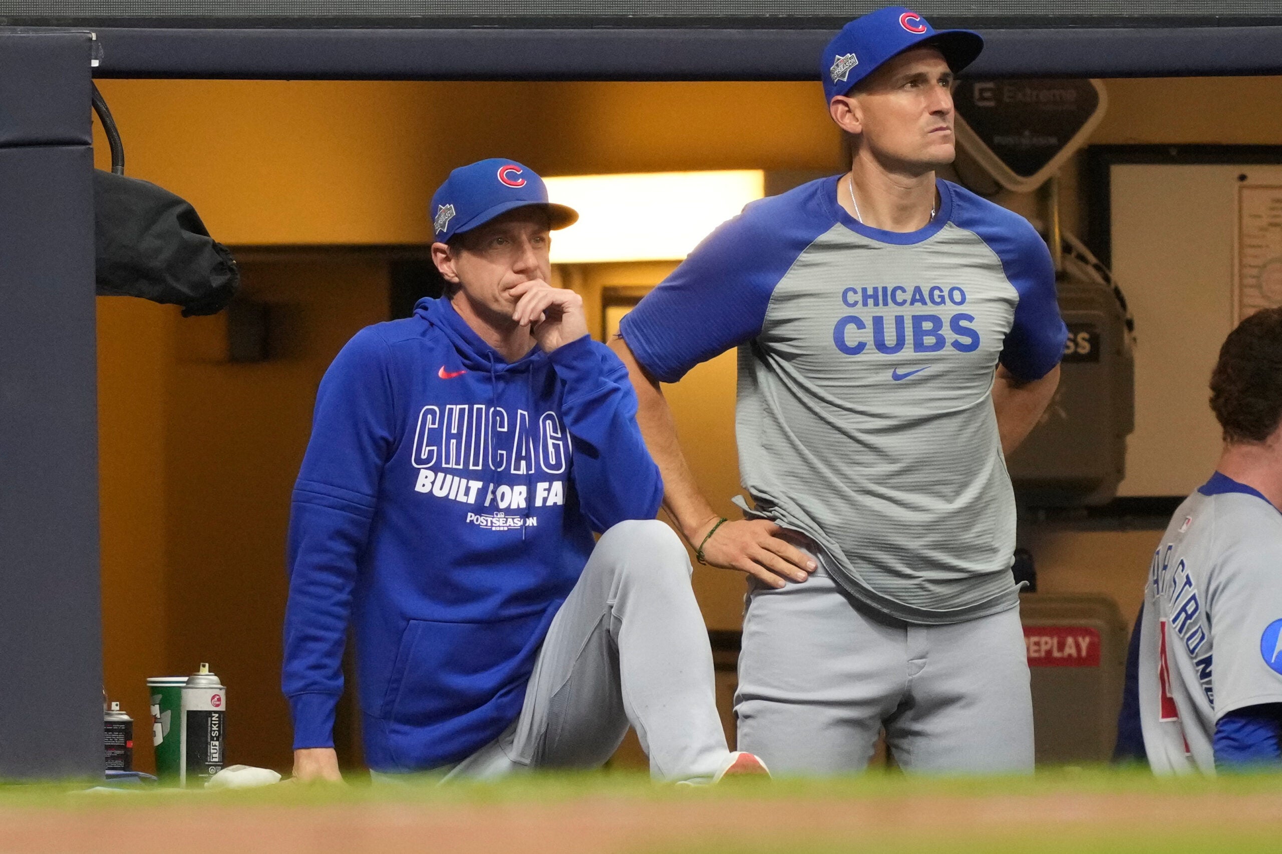 Two Chicago Cubs staff members in team apparel watch a game intently from the dugout, one standing with hands on hips and the other seated with a hand on his face.