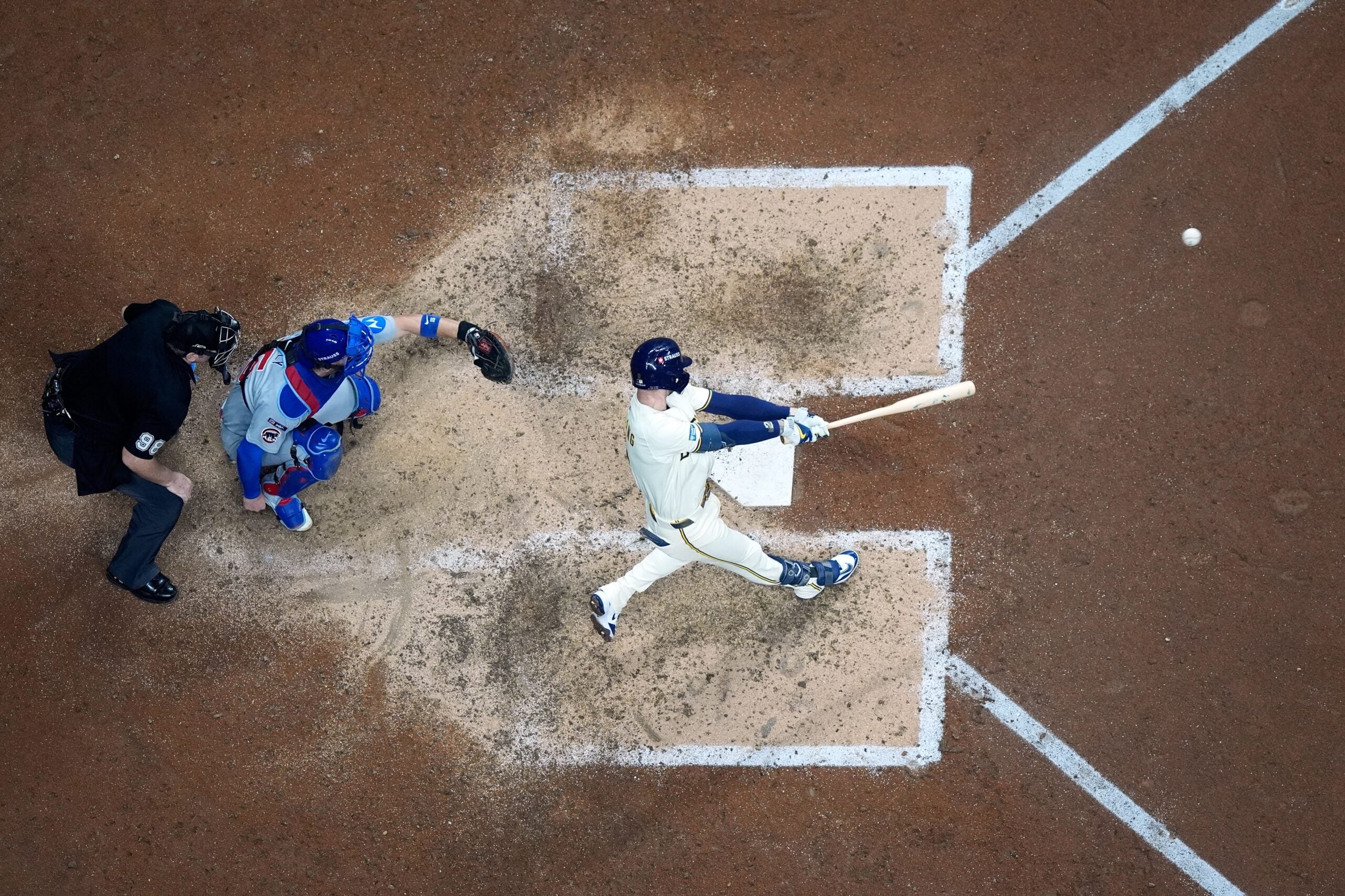 A baseball batter swings at a pitch as the catcher and umpire look on from behind home plate, viewed from above.