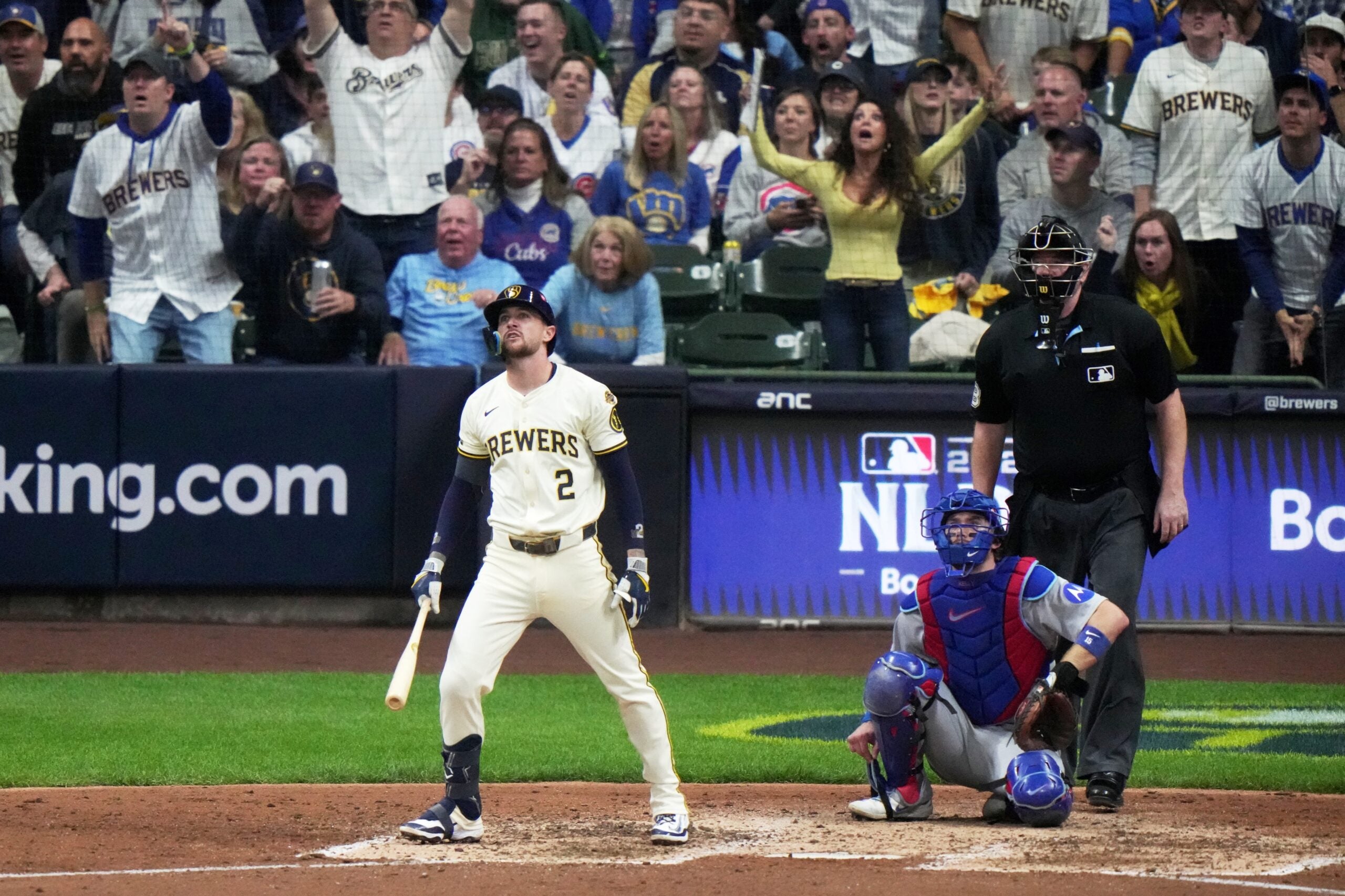A Milwaukee Brewers baseball player watches the ball after hitting, while fans and players react from the stands and dugout during a game. An umpire and catcher are behind him.