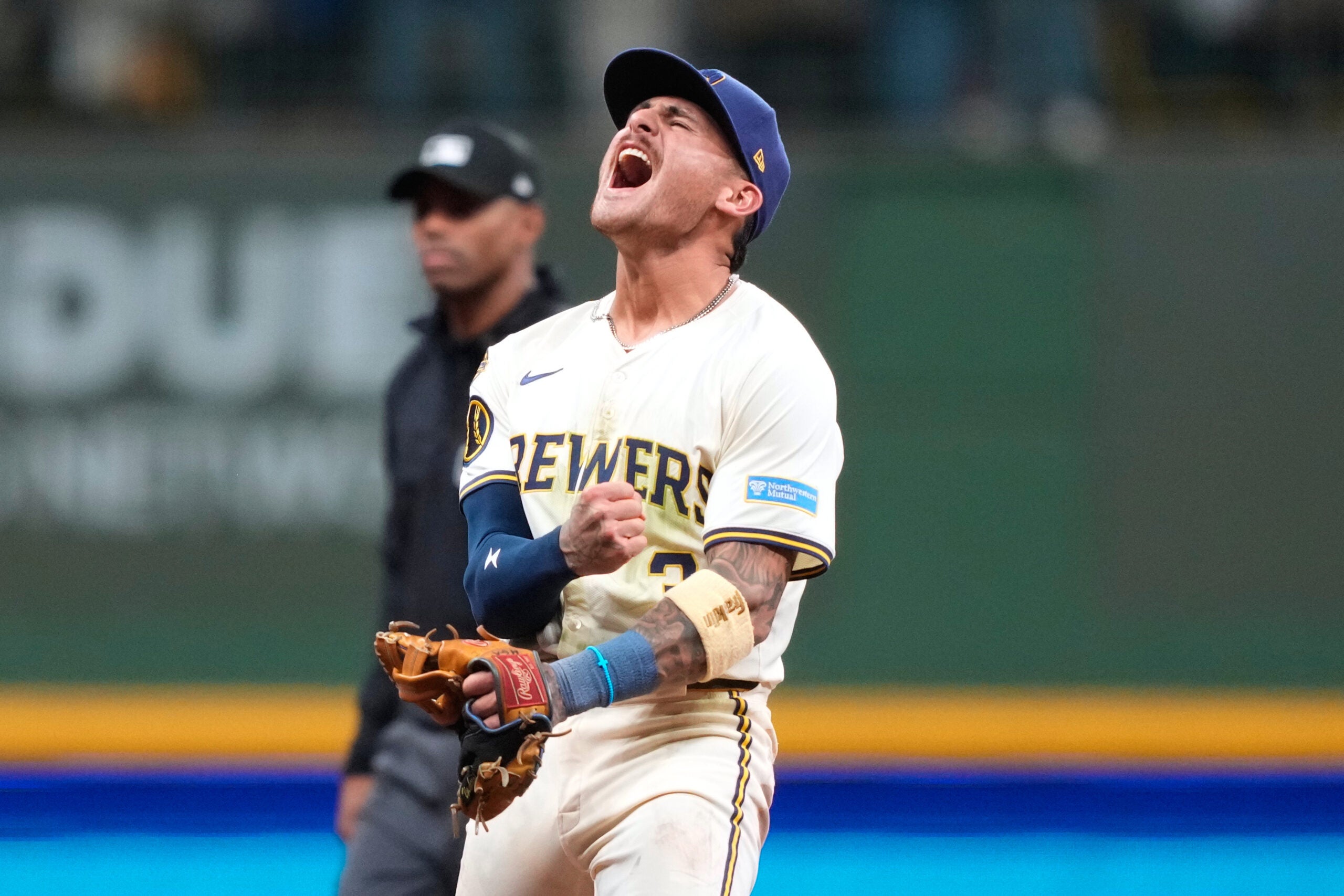 A Milwaukee Brewers baseball player in uniform yells in celebration on the field, clenching his fist, while an umpire stands in the background.