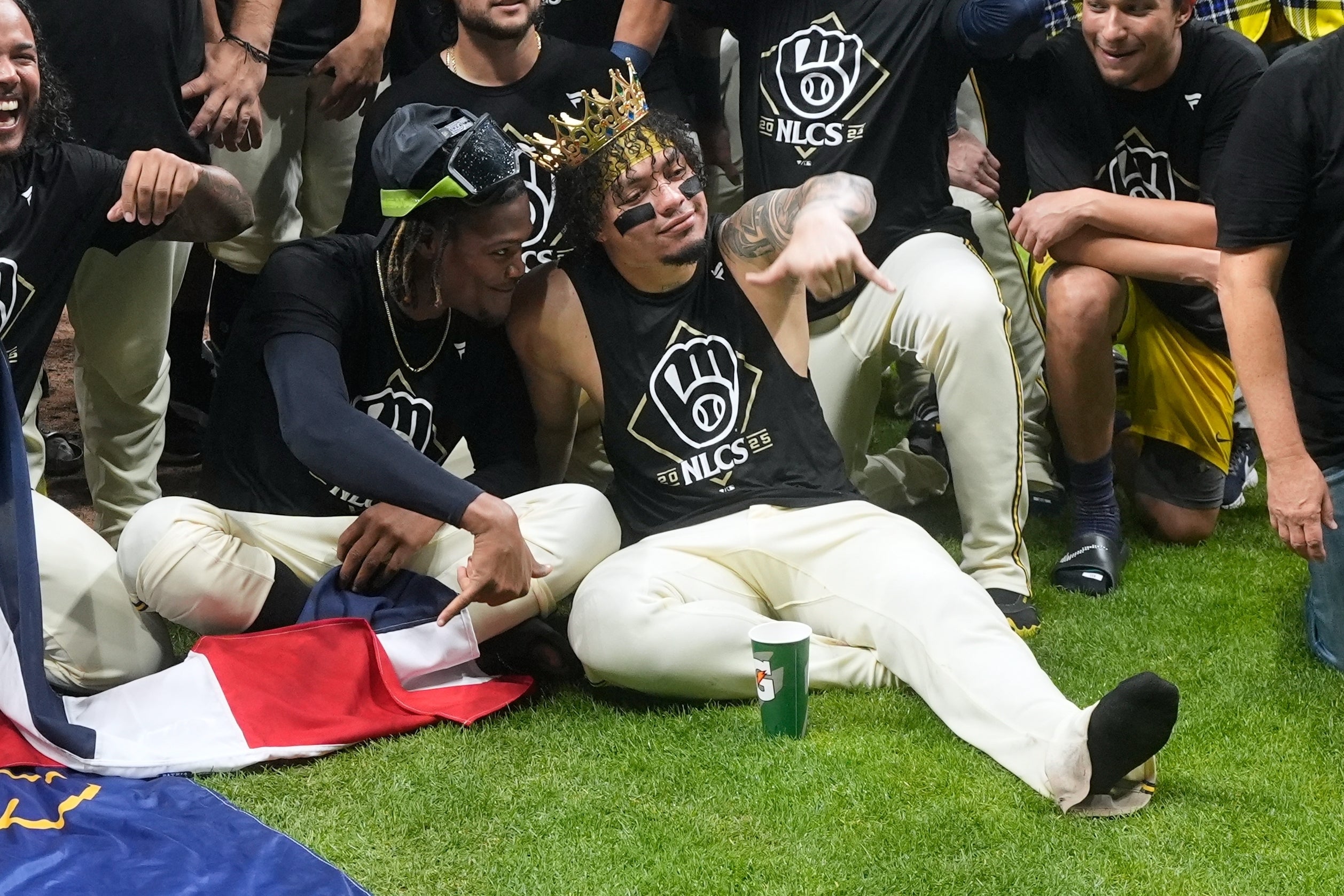 Baseball players wearing matching NLCS shirts sit on grass; one wears a crown and sunglasses, others are smiling and posing for a celebratory team photo.