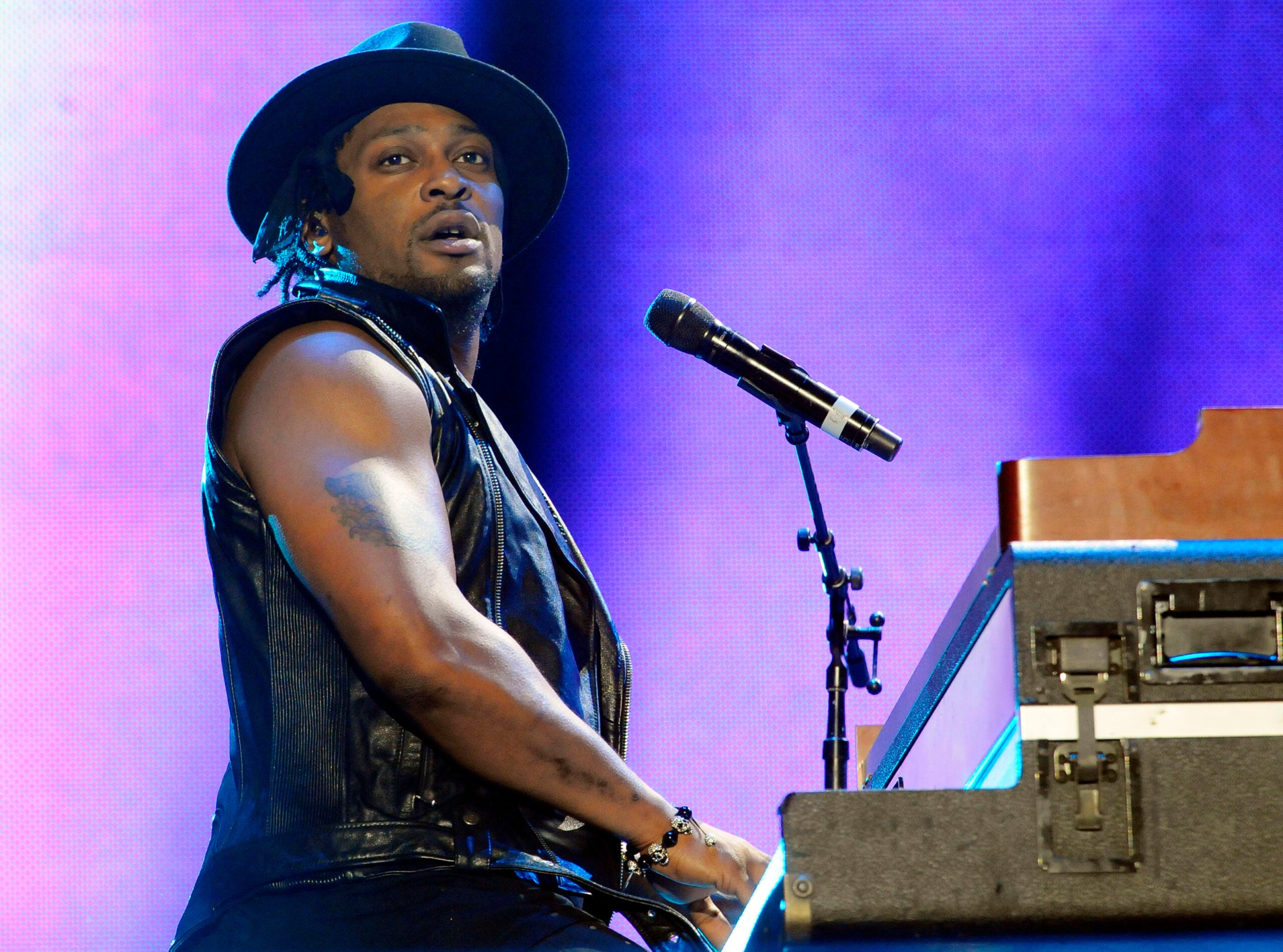 A musician wearing a black hat and sleeveless vest plays the keyboard and sings into a microphone on stage with purple lighting in the background.
