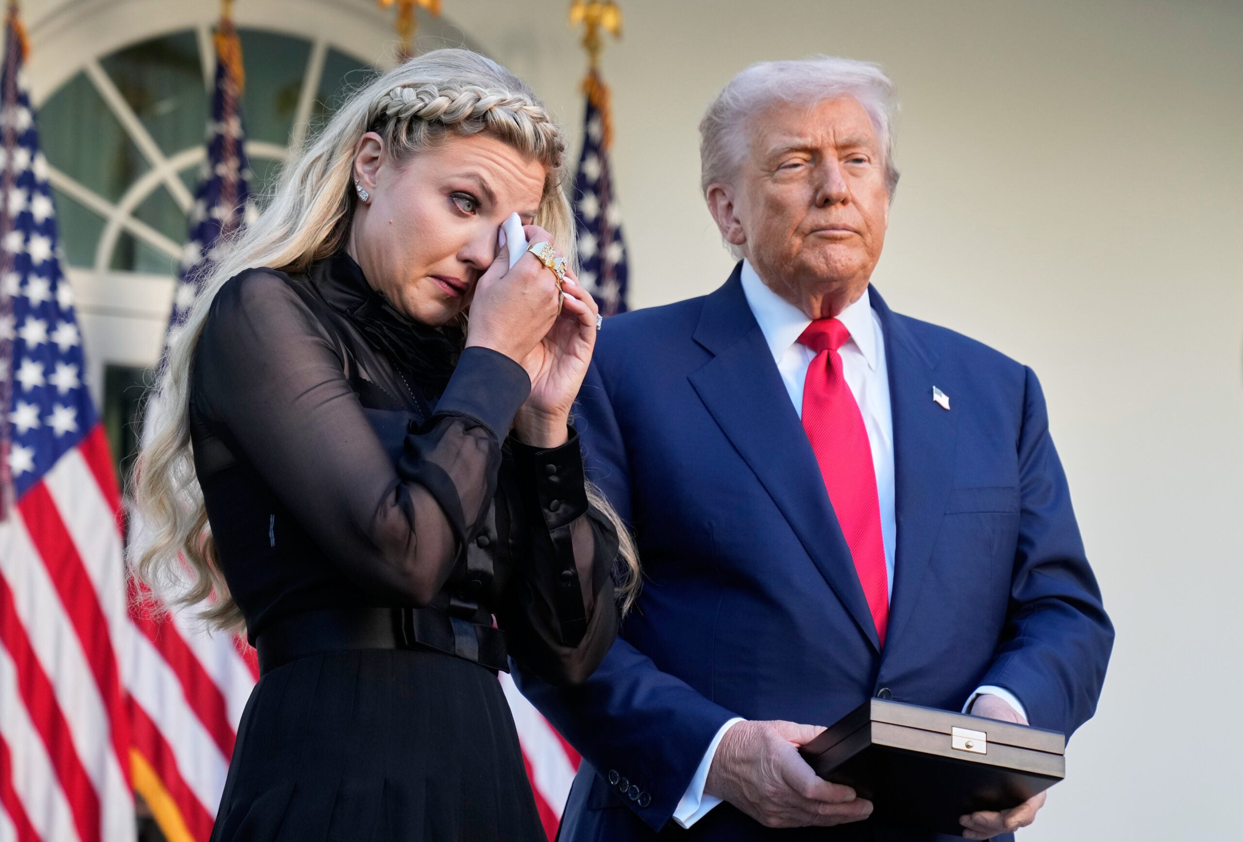 A woman wipes her eyes with a tissue while standing beside a man in a suit holding a box, with American flags in the background.