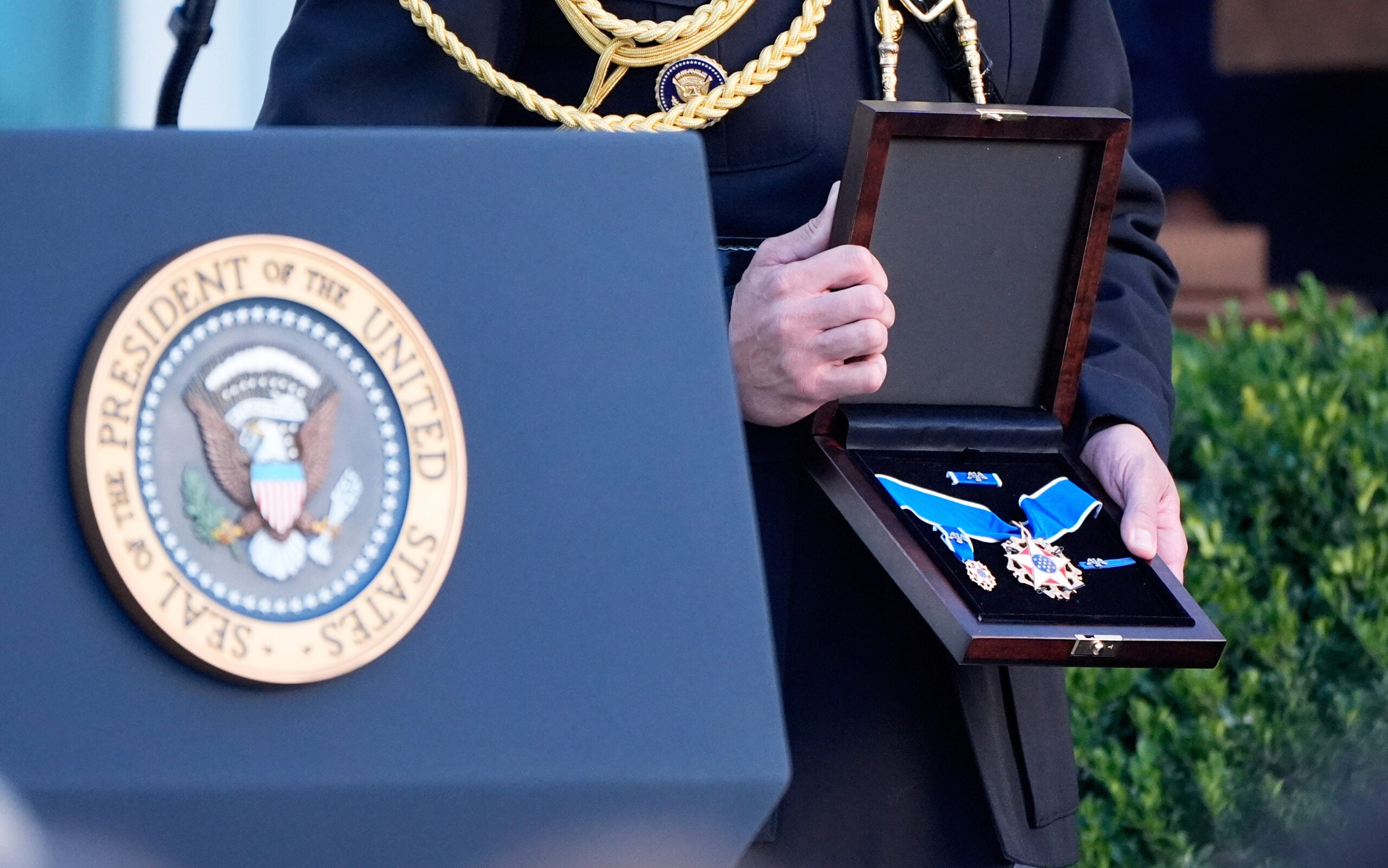 A person stands behind a podium with the U.S. presidential seal, holding an open box containing the Presidential Medal of Freedom.