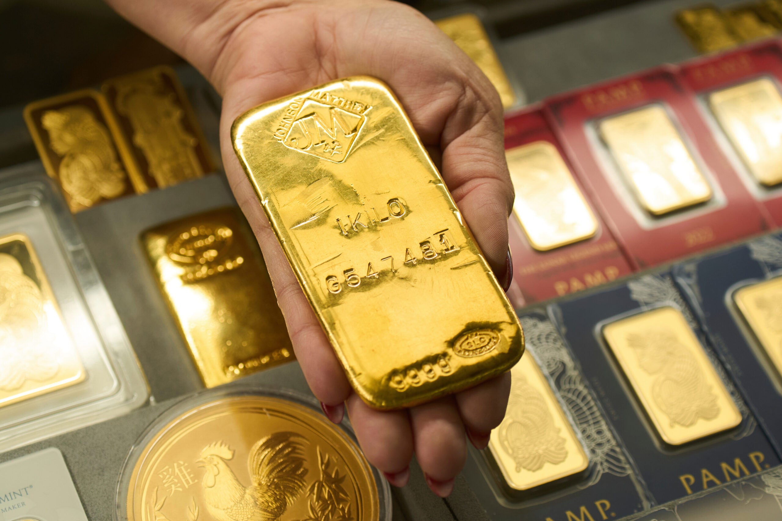 A hand holds a large gold bar above a display of various other gold bars and coins in protective cases.