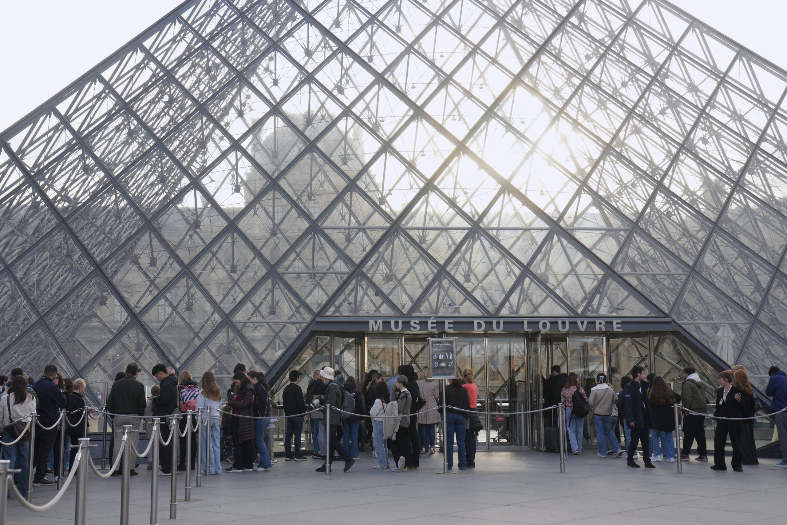 People wait in line outside the glass pyramid entrance of the Louvre Museum in Paris, with sunlight shining through the structure.