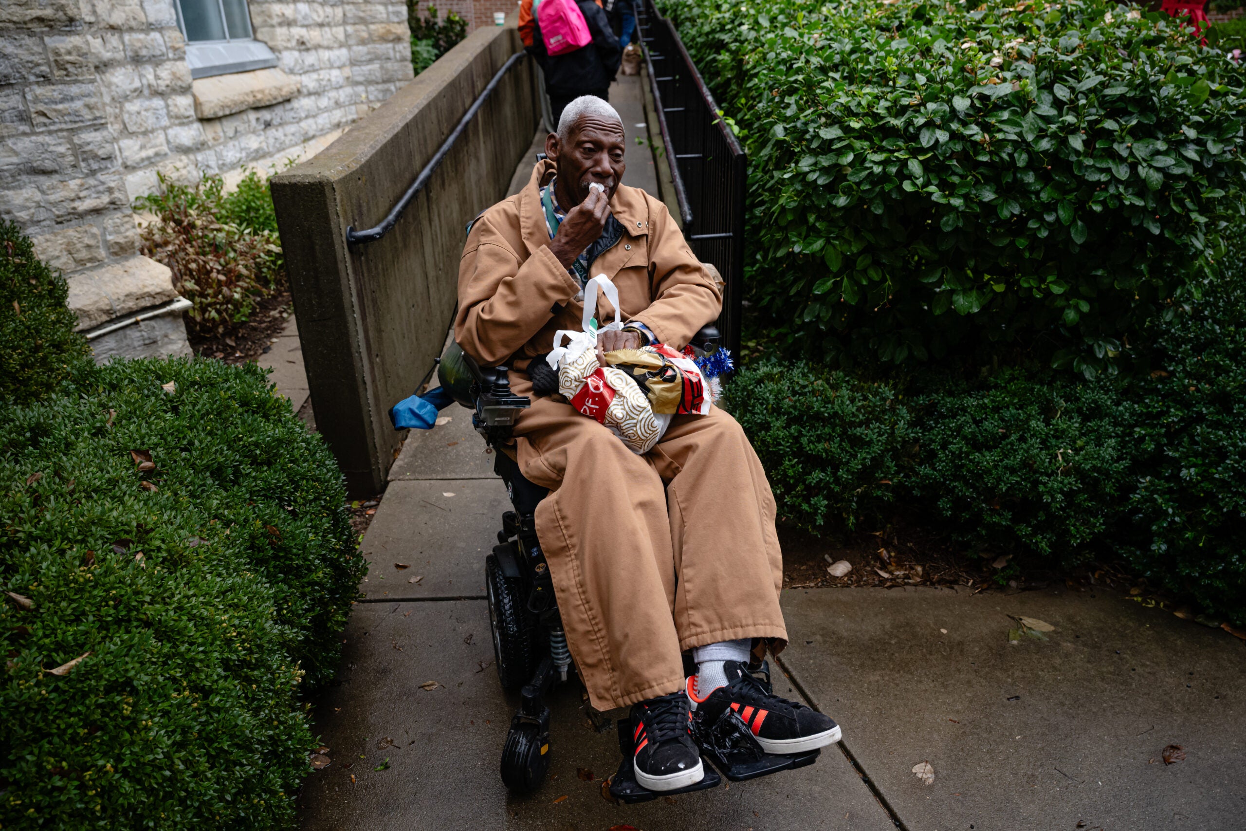 An elderly man in a tan coat sits in a motorized wheelchair on a sidewalk, holding plastic bags and using a cellphone. Bushes and a stone building are nearby.