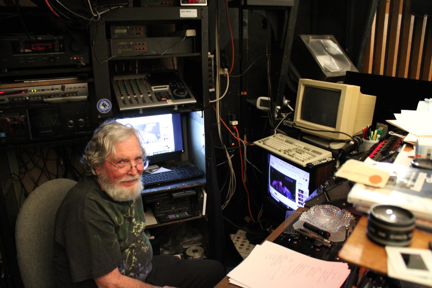 An older man with a white beard sits in a cluttered room filled with vintage computer and audio equipment, looking toward the camera.