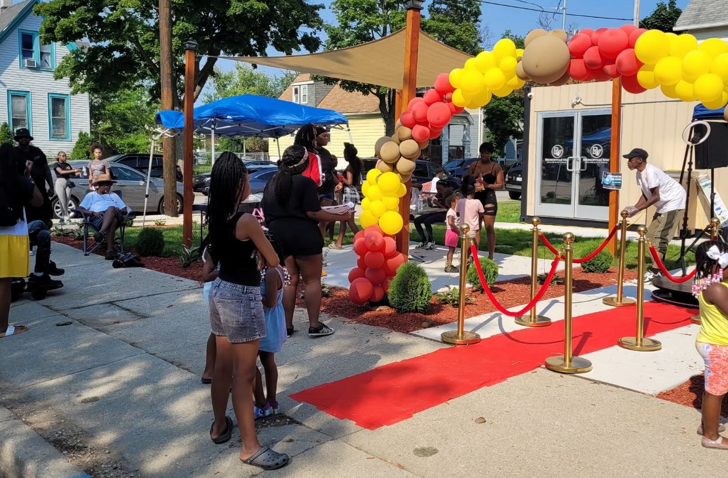 People gather outdoors near a red carpet and a balloon archway, with some standing in line and others sitting, on a sunny day in a residential neighborhood.