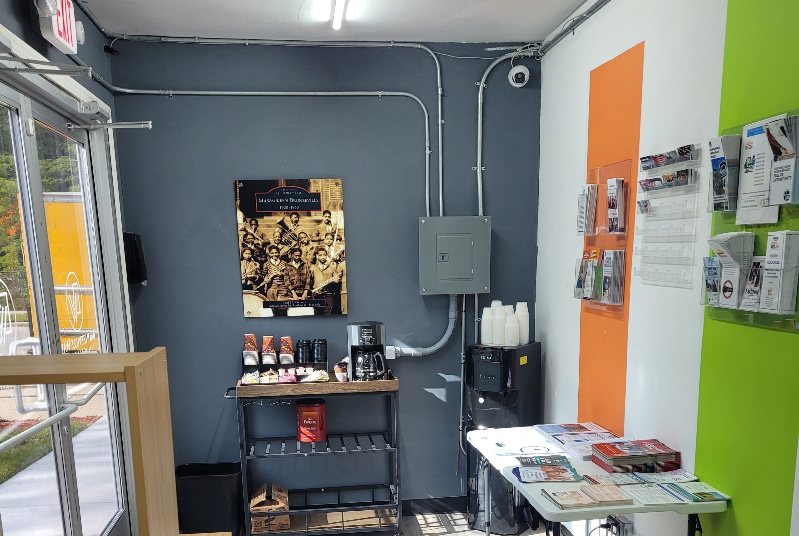 A small waiting area with brochures, a coffee station, paper cups, a wall calendar, and a black-and-white photo on the wall near a glass door.