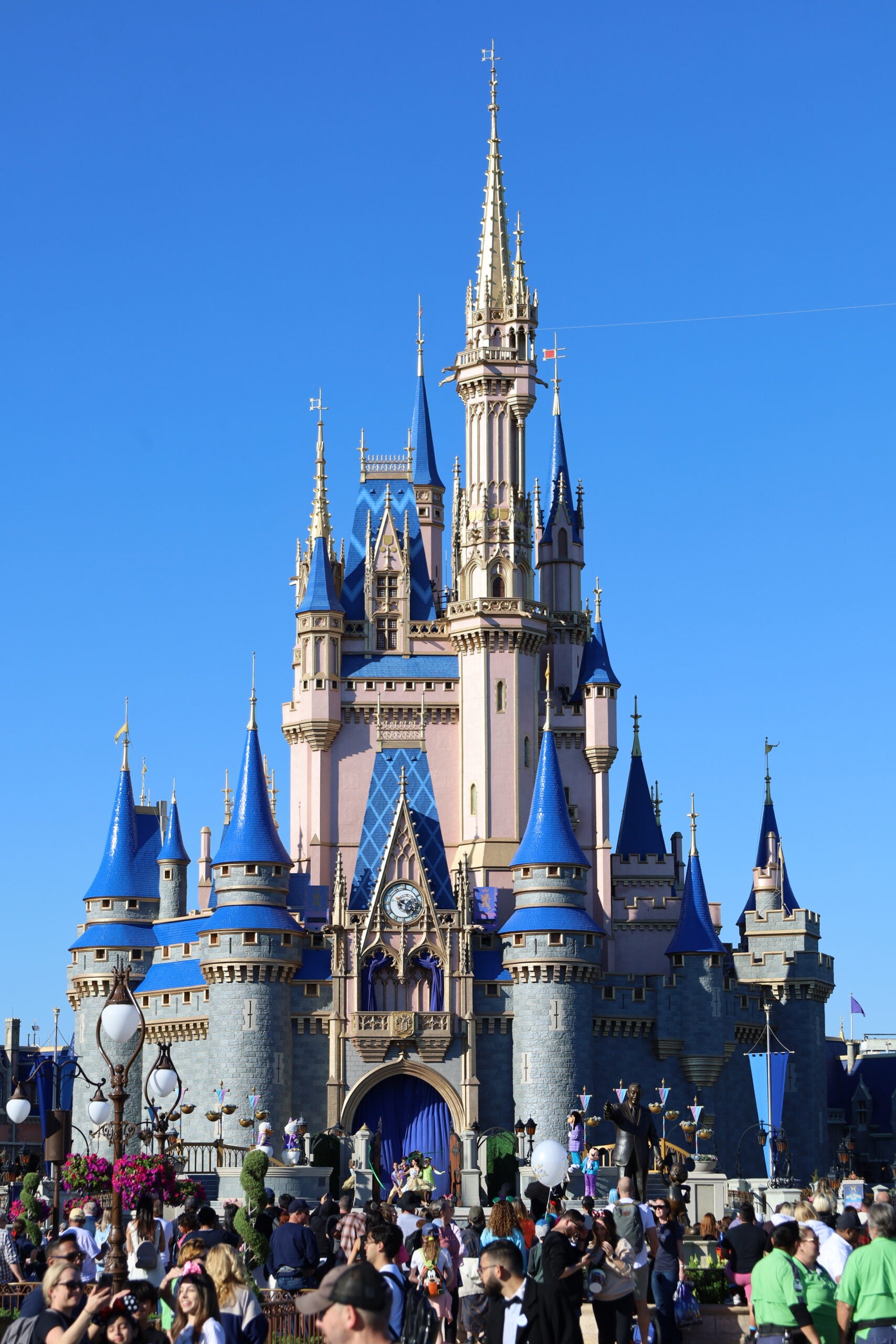 Crowds of people gather in front of a large, ornate castle with blue and gold spires, set against a clear blue sky.
