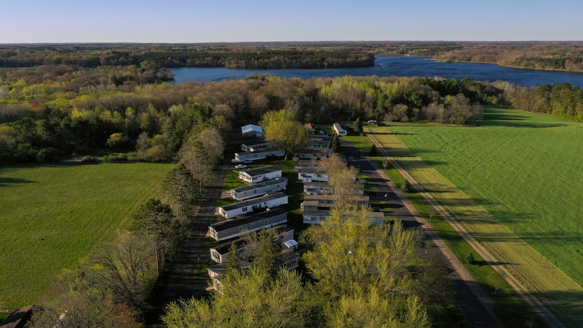 Aerial view of a small mobile home park surrounded by green fields and trees, with a lake and forested area in the background under a clear sky.