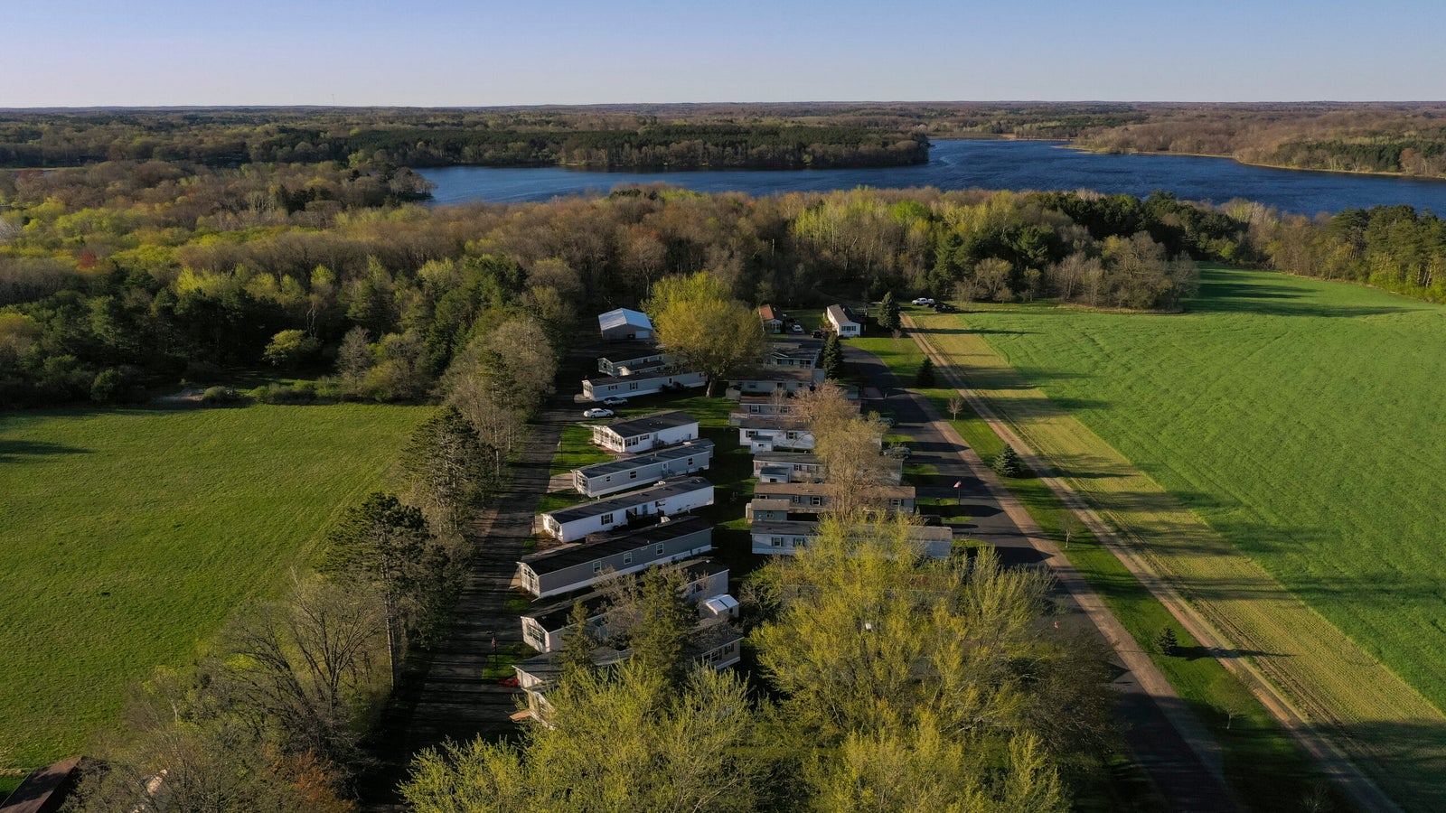 Aerial view of a small mobile home park surrounded by green fields and trees, with a lake and forested area in the background under a clear sky.