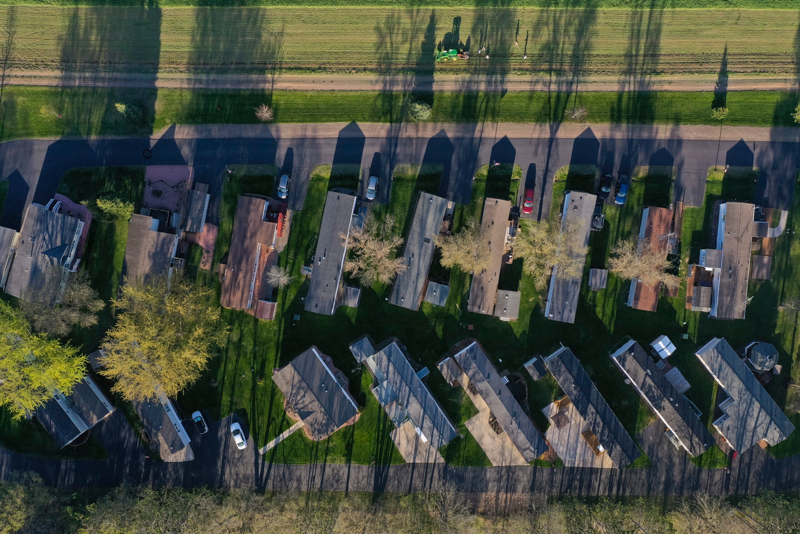 Aerial view of a row of mobile homes with parked cars, green lawns, and long tree shadows cast on a sunny day.