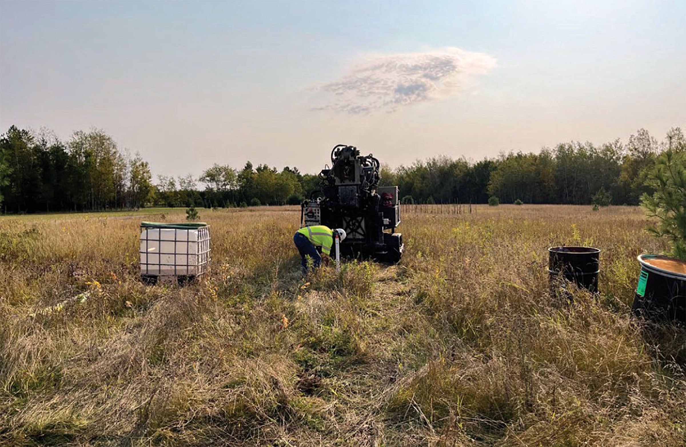 A worker in a safety vest bends near a large machine in a grassy field, with barrels and a large white water container nearby. Trees and a clear sky are in the background.