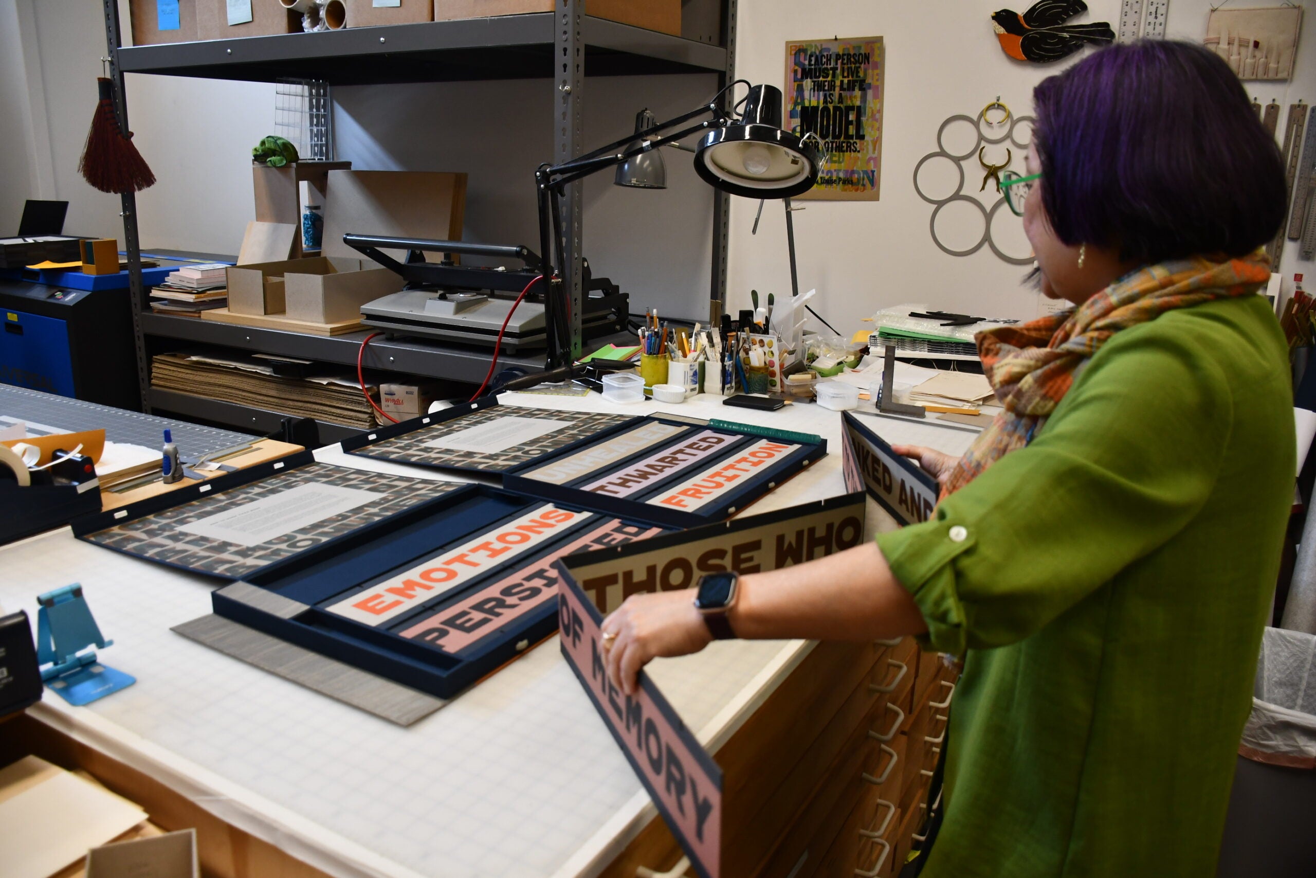 A person arranges framed text panels on a worktable in an art studio, with supplies and tools visible in the background.